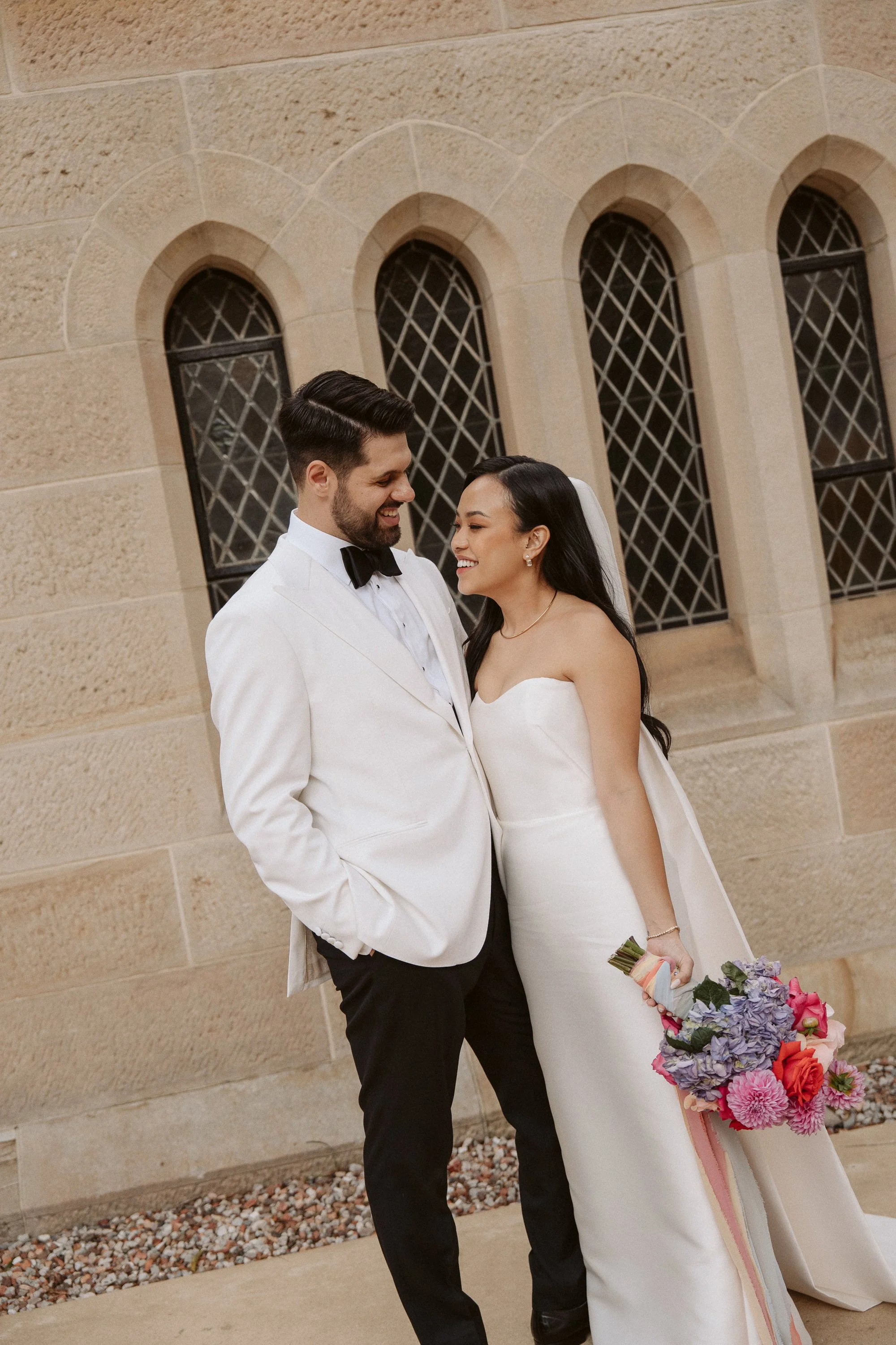 A bride and groom smiling at each other outside a church, with the bride holding a bouquet of pink and purple flowers.