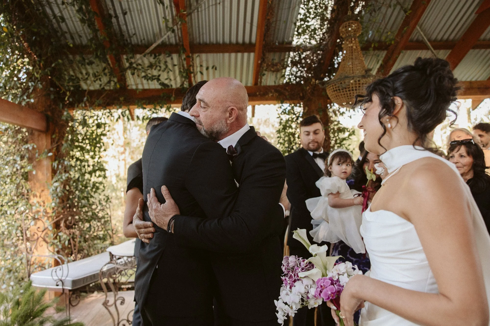 Two men are hugging at a wedding ceremony with a bride holding a bouquet of flowers and a young girl in a white dress looking on. Other guests are in the background.