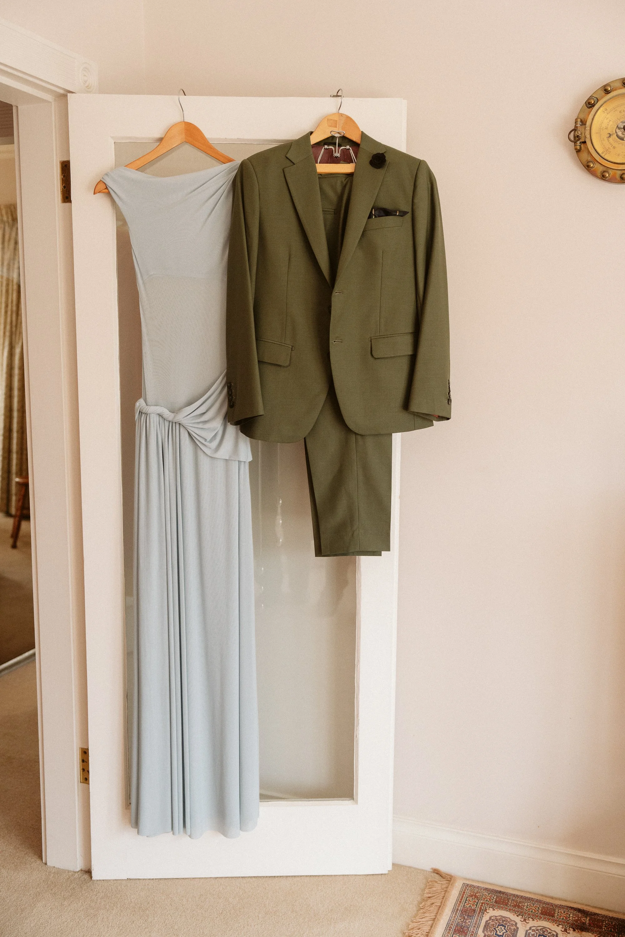 A white door with an olive green suit hanging on it, and a light blue dress hanging on a hanger next to the suit.