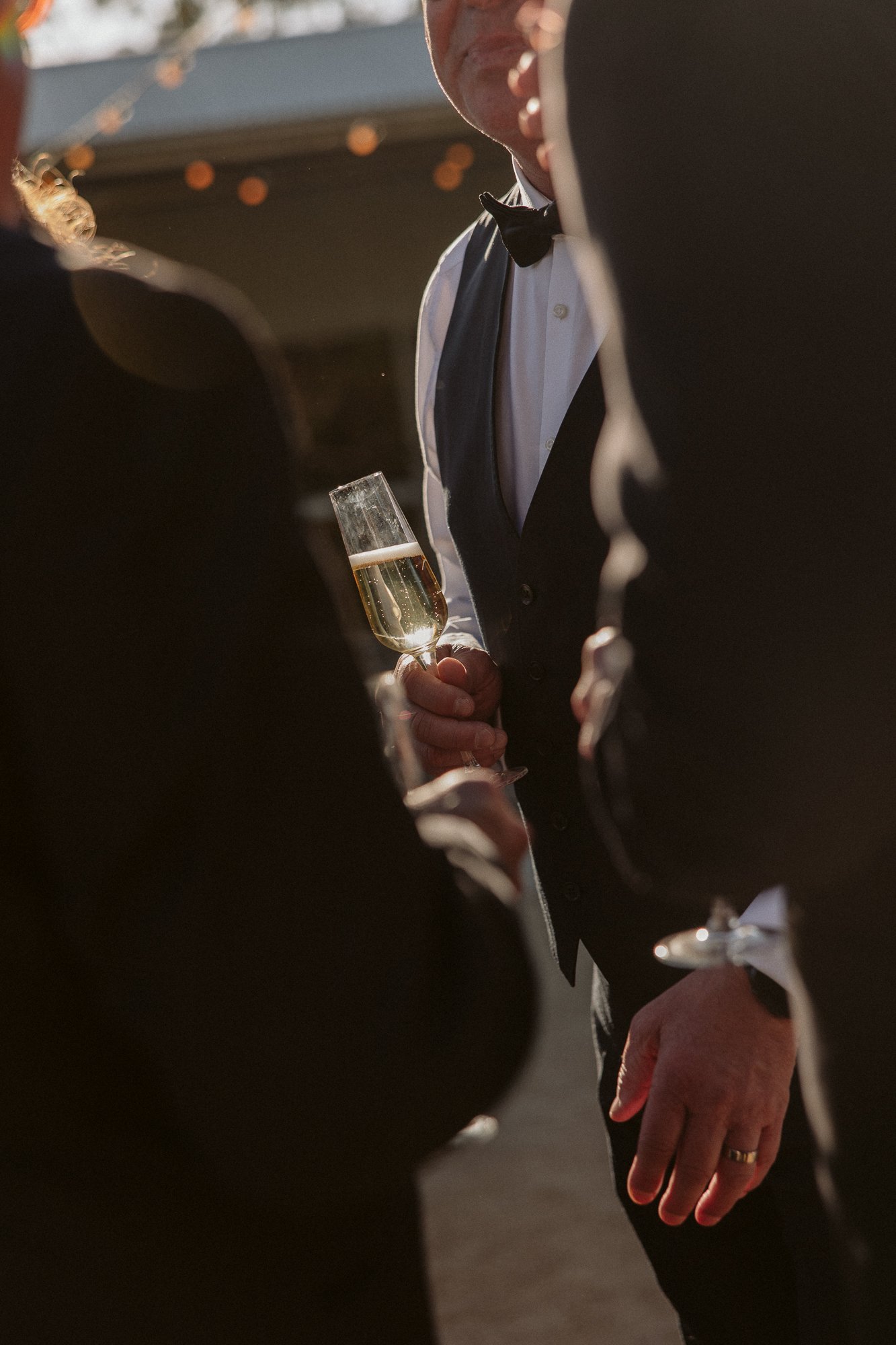 People wearing formal attire, including tuxedos and dresses, gathered outdoors at a social event during the evening, with one person holding a glass of champagne.