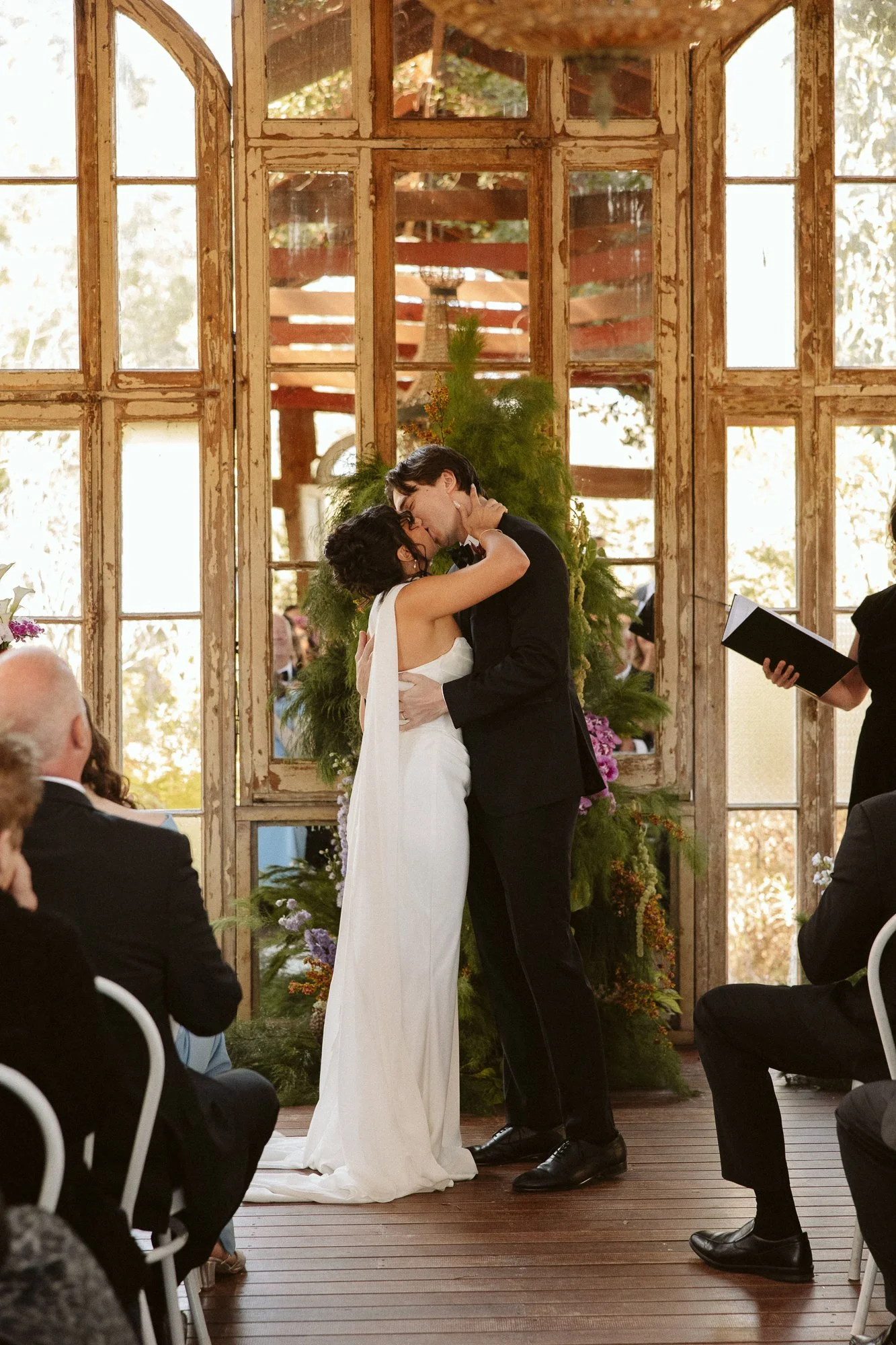 A bride and groom sharing a kiss during their wedding ceremony, surrounded by seated guests. The ceremony is set indoors in front of rustic, vintage windows with greenery and flowers as decoration.