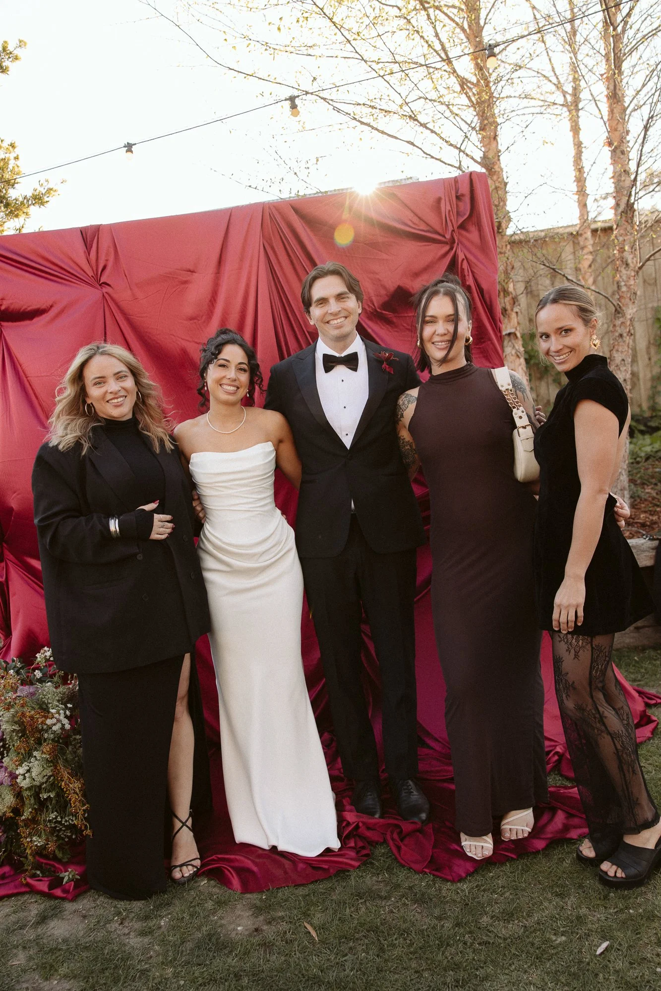 Group of five people dressed in formal attire at an outdoor event, with a red draped backdrop and trees in the background.
