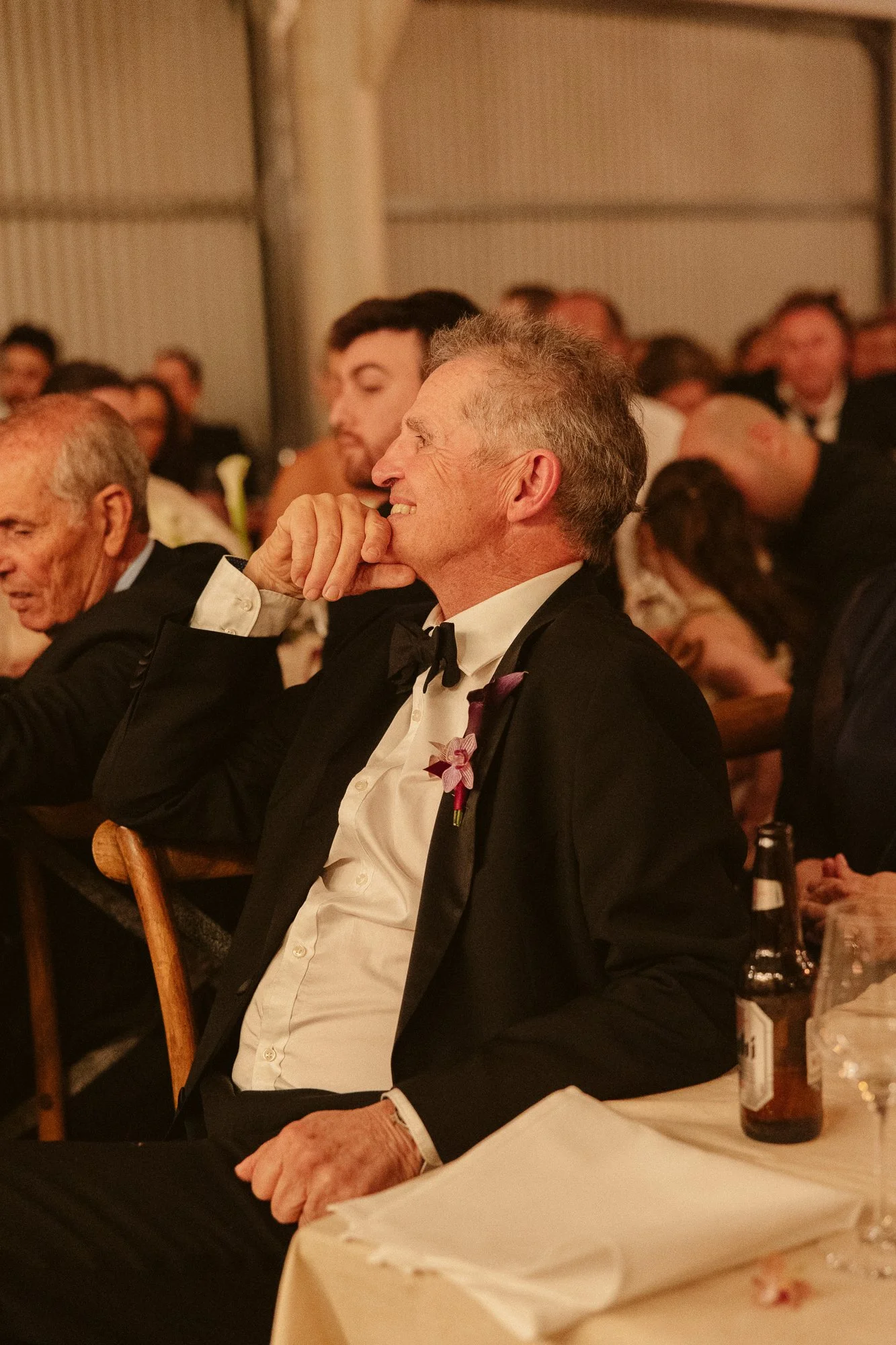 A man in a tuxedo and bow tie is smiling while seated at a formal event.