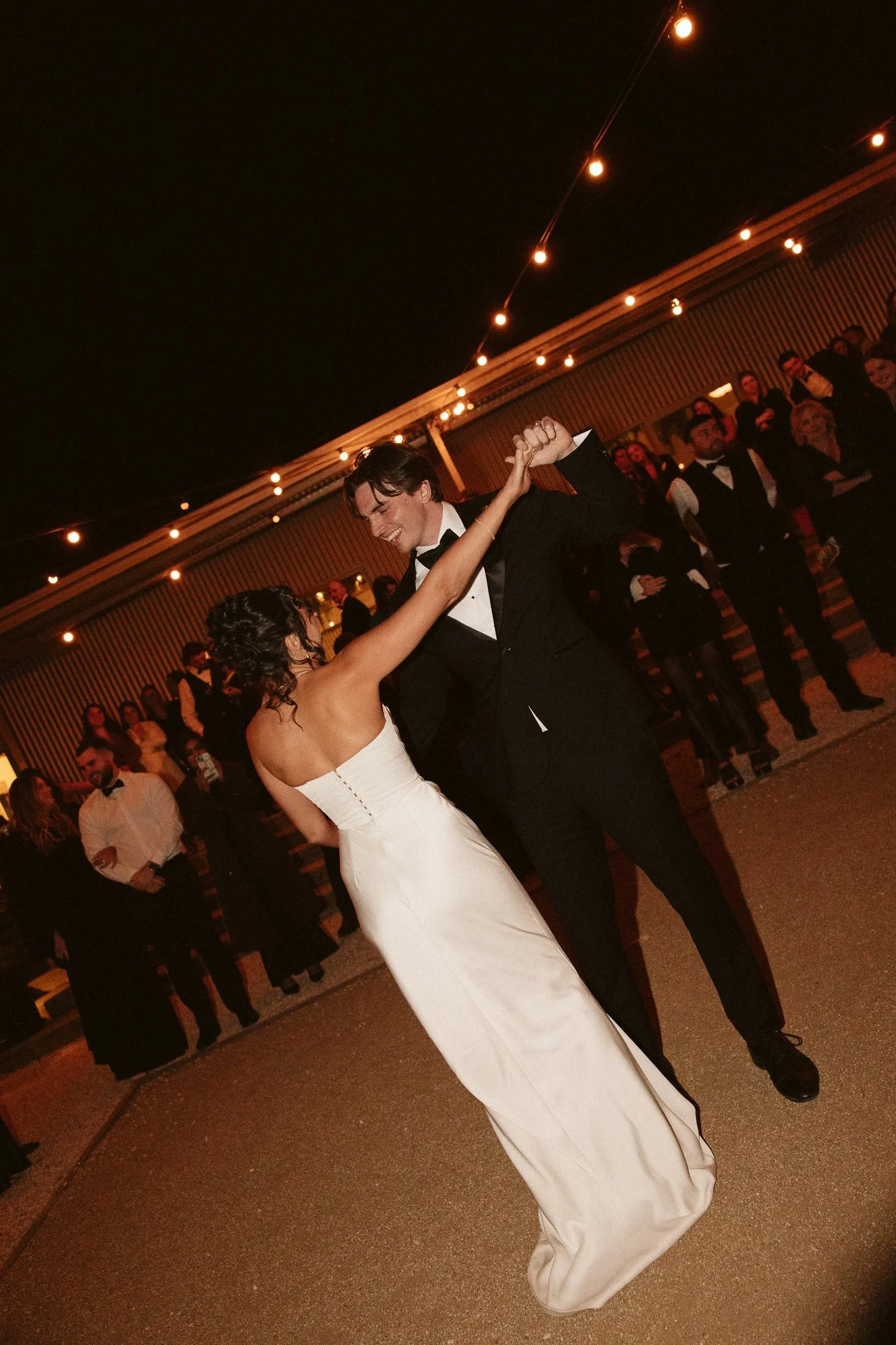 A couple dancing at their wedding reception, with guests watching in the background under string lights at night.