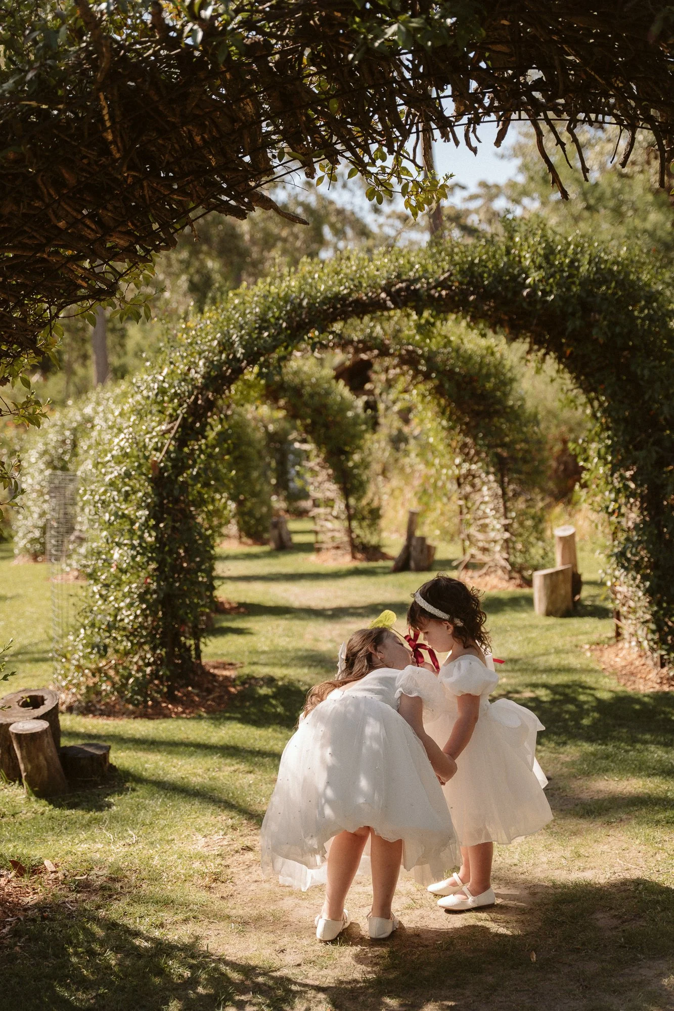 Two young girls in white dresses sharing a kiss under a leafy archway in a garden.