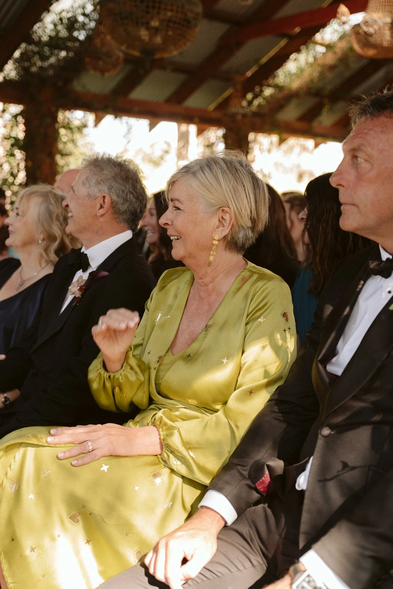A group of elegantly dressed people seated at an event, including a woman in a shiny yellow-green dress laughing and a man in a tuxedo.