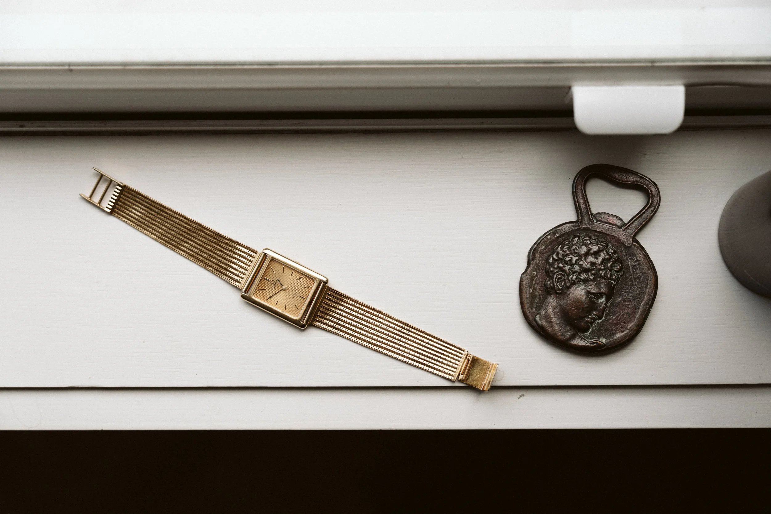A gold wristwatch with a mesh band and a square face, and a bronze-colored medallion with a profile of a woman's face and a lock-shaped loop at the top, placed on a white surface near a window.