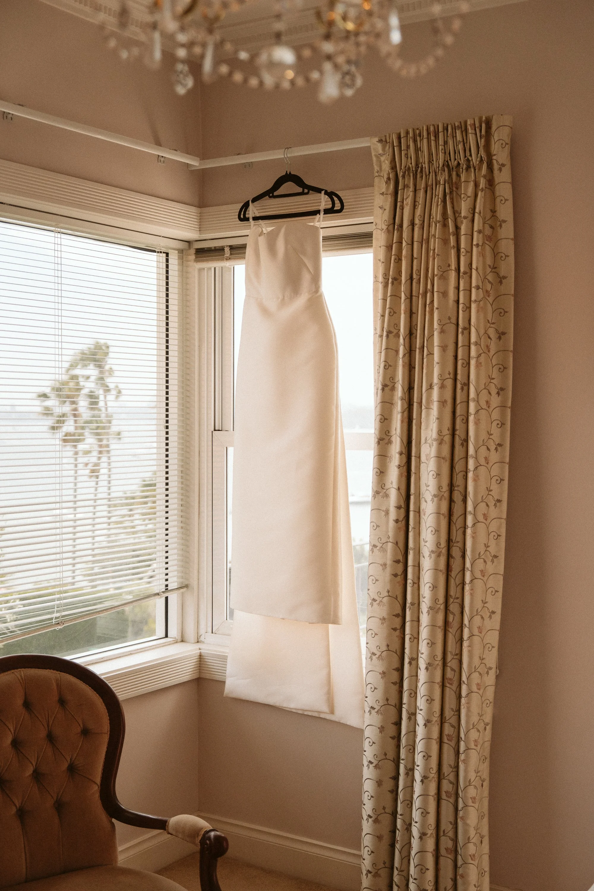 A white wedding dress hanging on a black hanger from a curtain rod, with beige curtains and a window with blinds in the background.