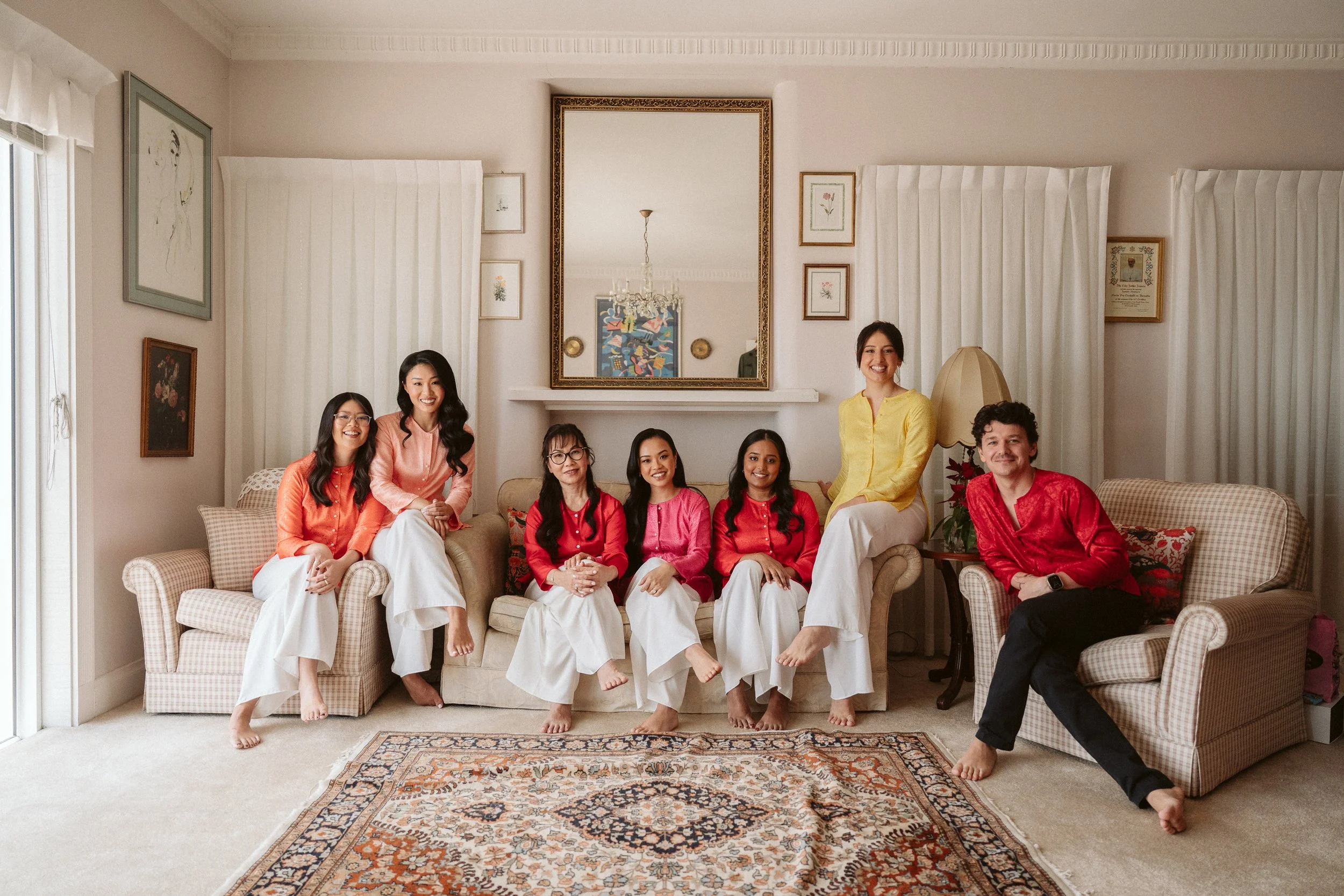 Group of seven women and one man sitting and standing on a sofa in a living room, smiling at the camera. The women are wearing pink, red, and yellow blouses with white pants, while the man is wearing a red shirt with black pants. The room has framed 