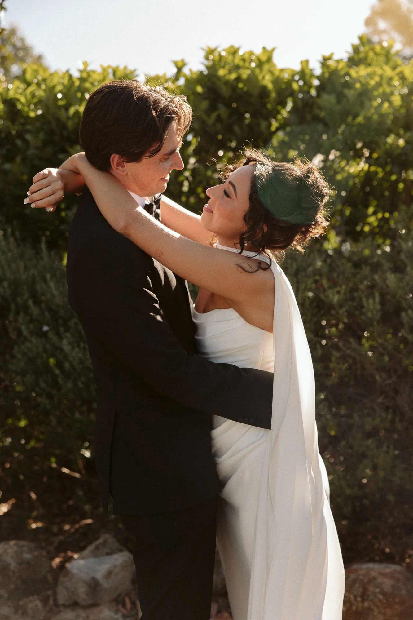 A couple in wedding attire embracing outdoors in a lush green setting during sunset.