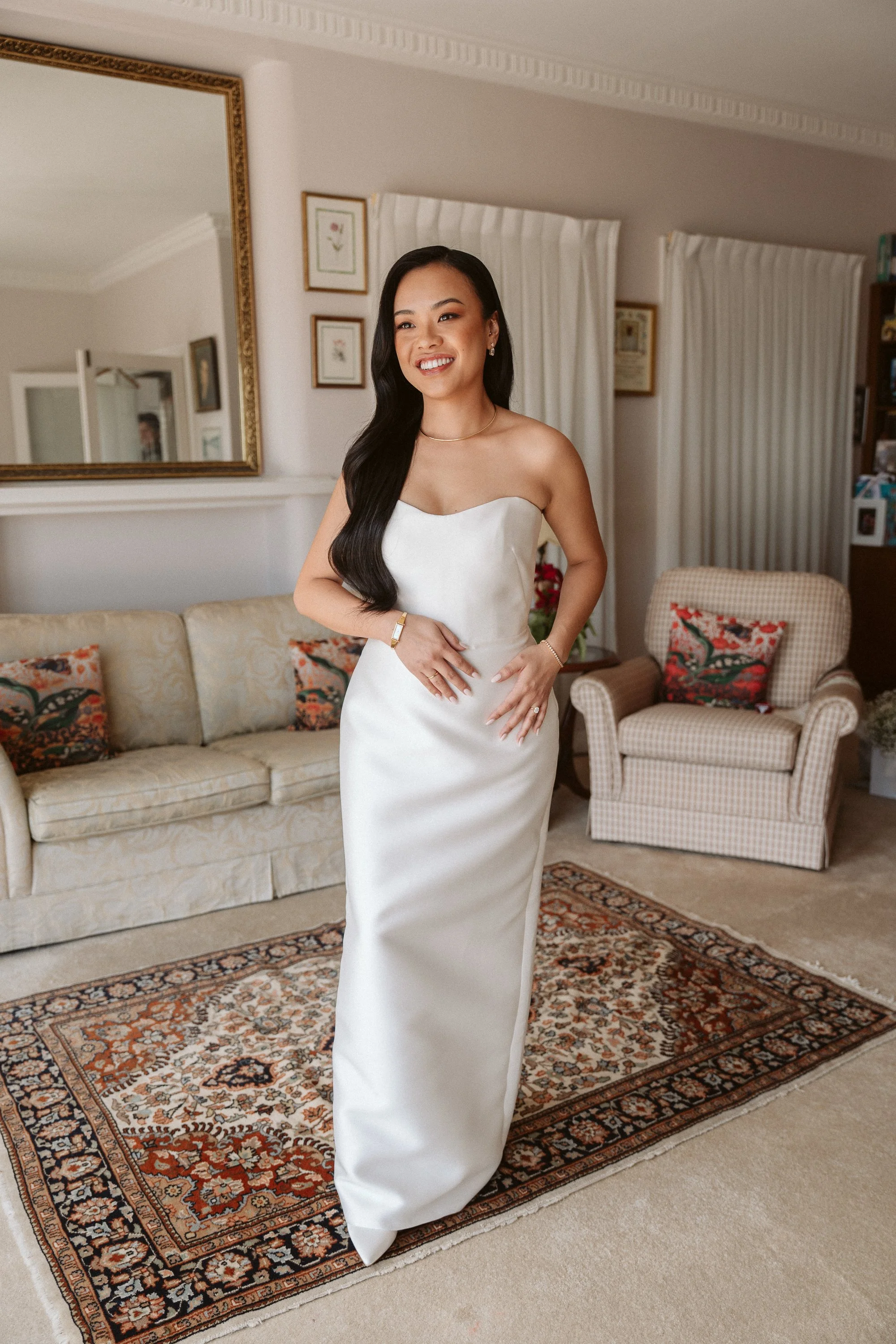 A woman with long black hair wearing a white strapless dress standing in a living room, smiling, with a patterned rug, sofa, and armchair in the background.
