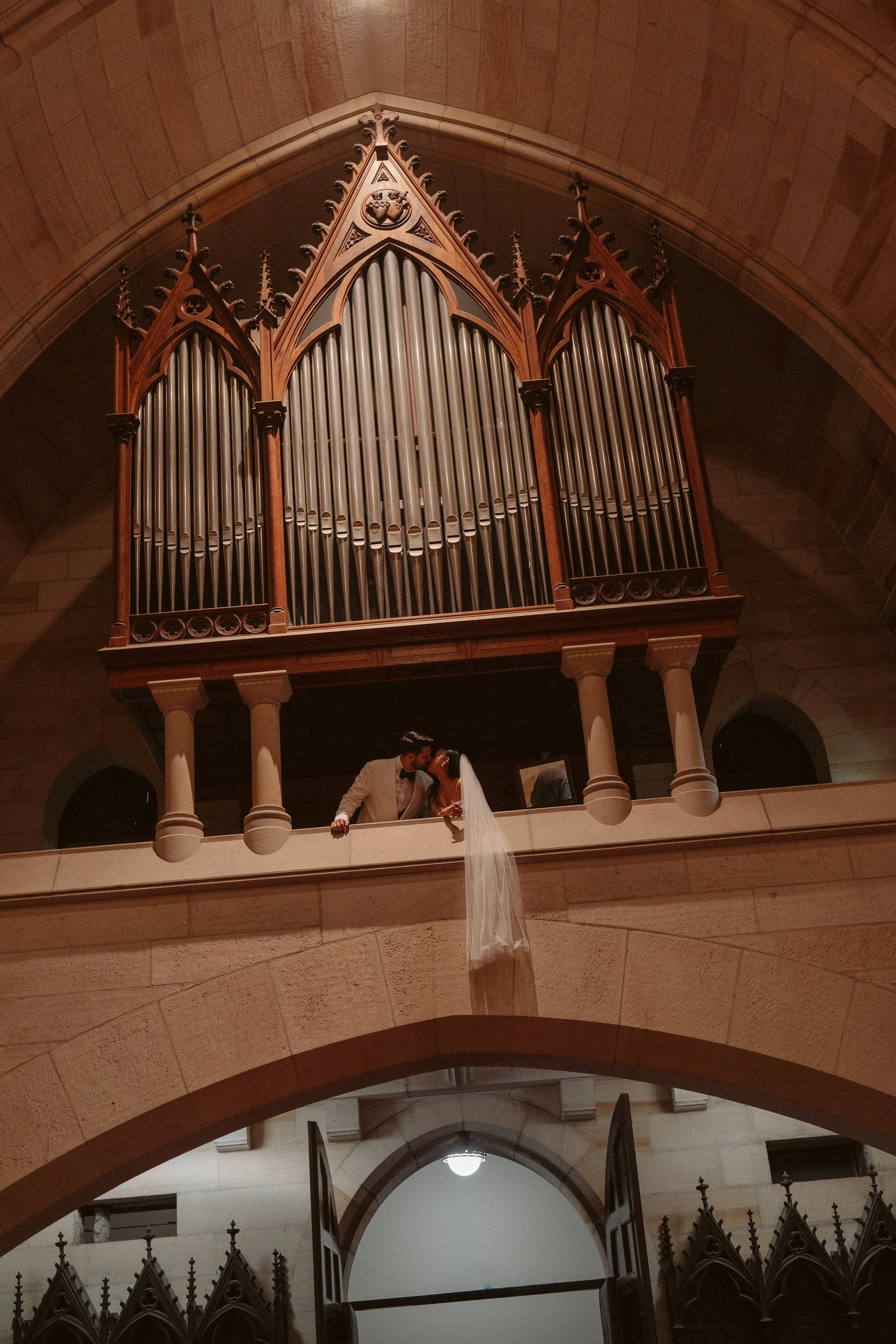 A bride and groom kissing on a balcony with a large pipe organ above them in a church.