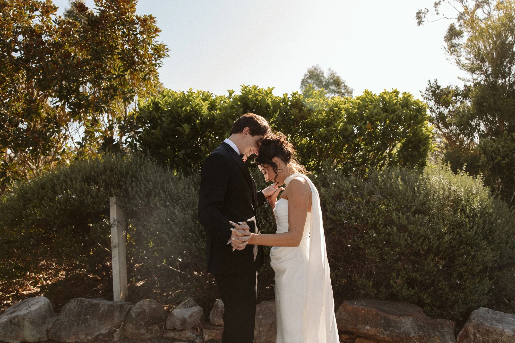 A couple in formal attire holding hands, smiling, outdoors near bushes and rocks, during what appears to be a wedding or special occasion.