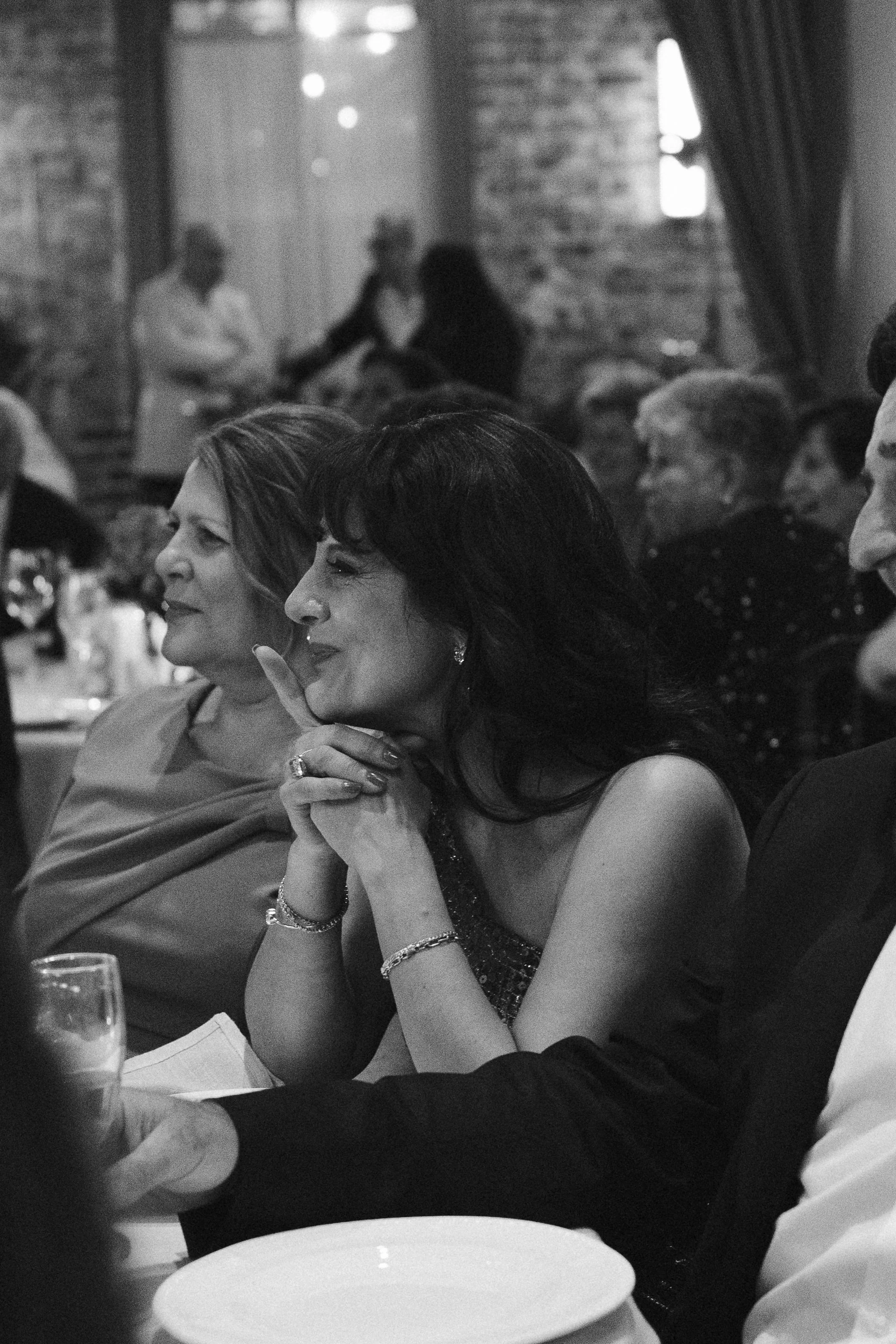 A woman with dark hair, wearing a dress, sitting at a table, smiling, with her hands clasped under her chin, during a social event in a dimly lit room with other people in the background.