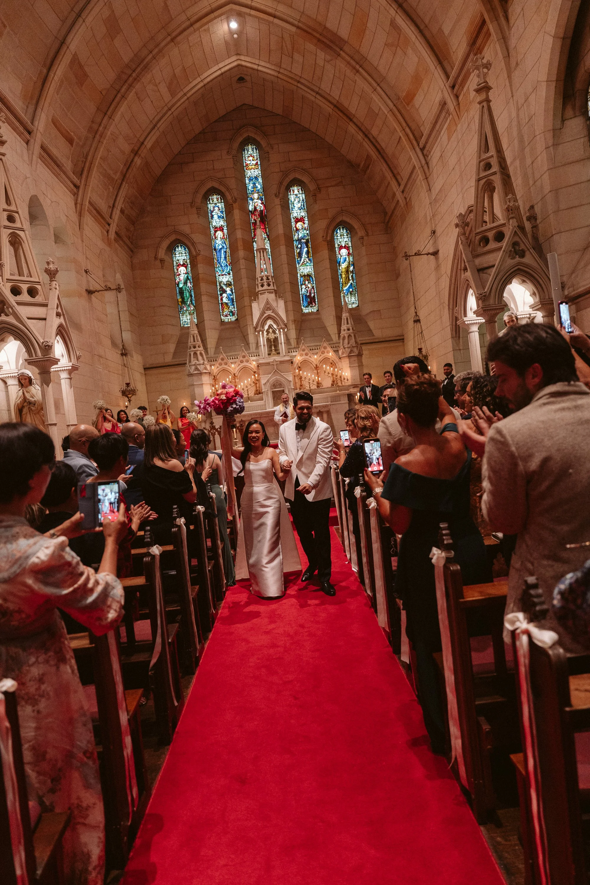 A bride and groom walking down the aisle after their wedding ceremony inside a church, surrounded by guests taking photos and clapping.