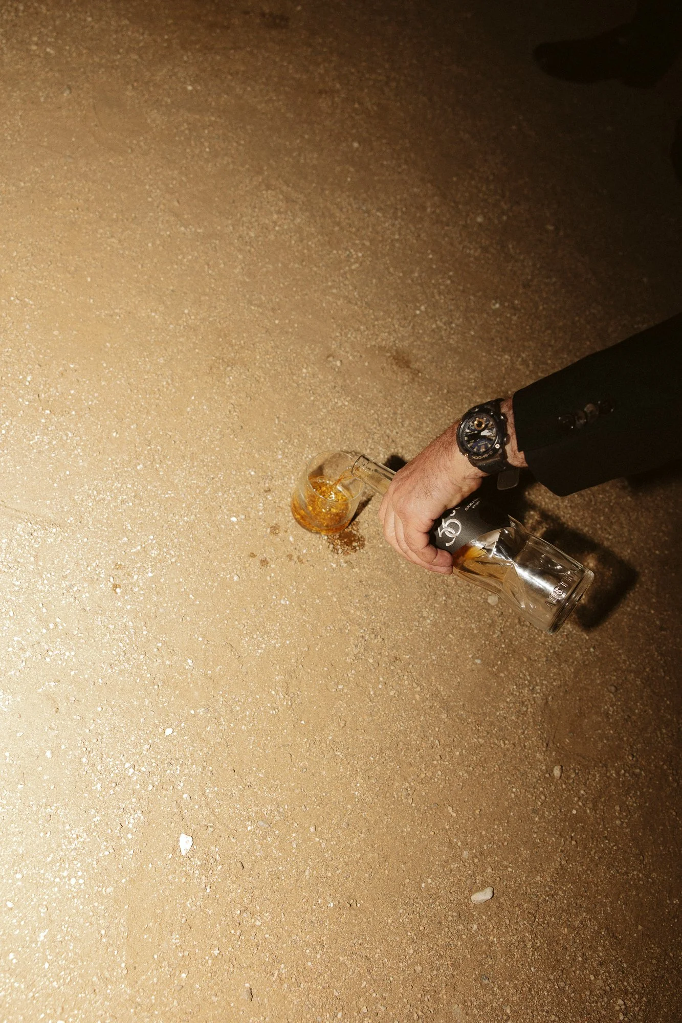 A person pouring whiskey from a bottle into a glass on sandy ground.