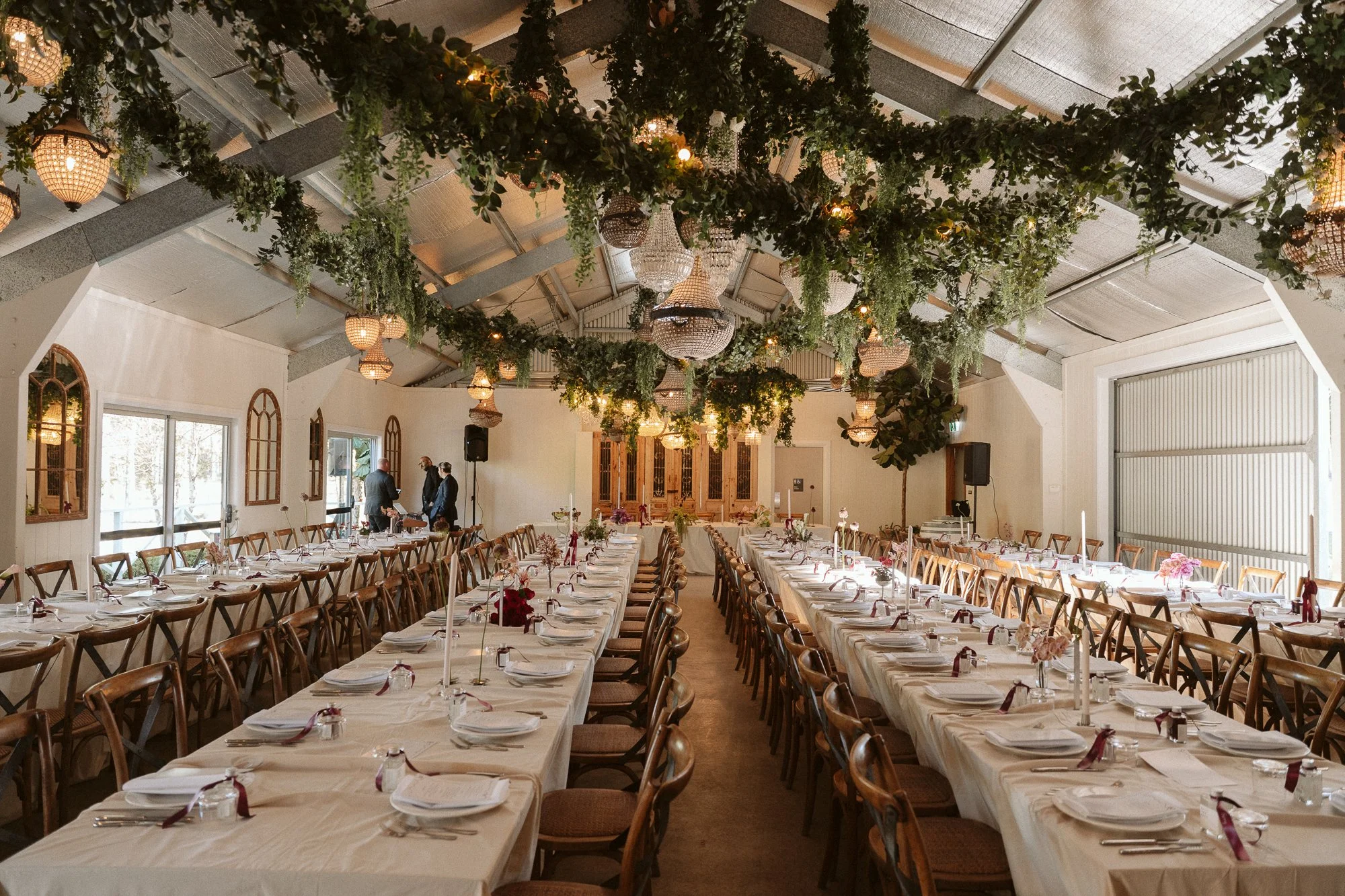 A decorated banquet hall with long tables set for a meal, featuring white tablecloths, plates, silverware, and pink and red floral centerpieces, green foliage hanging from the ceiling, and chandeliers.