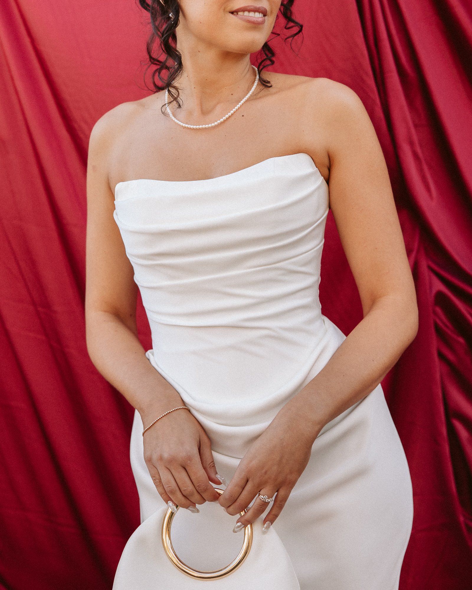 A woman in a white strapless dress holding a white circular handbag with gold rings, standing against a red curtain background.