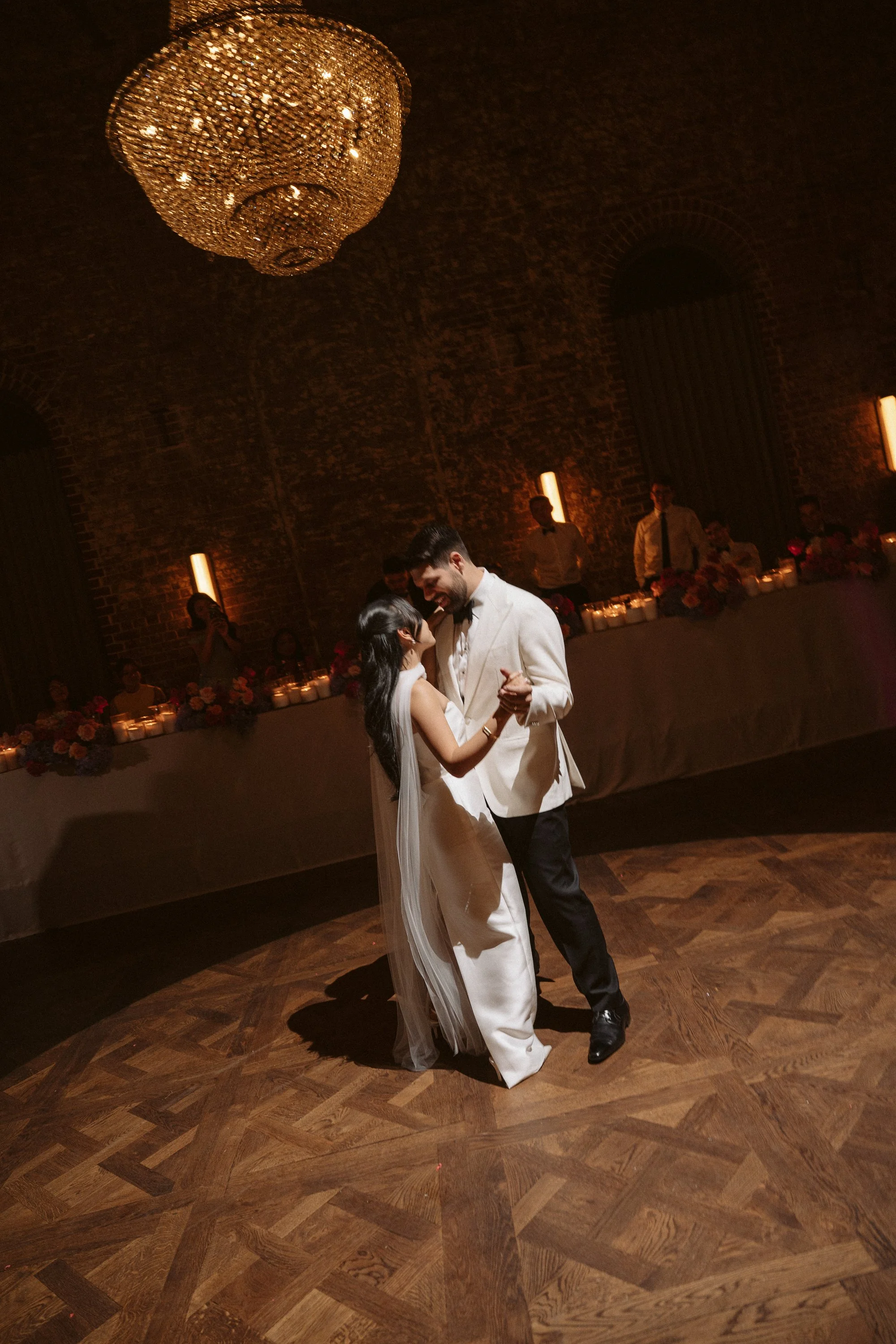 A couple dancing at their wedding reception, with guests watching in the background and a large chandelier overhead.