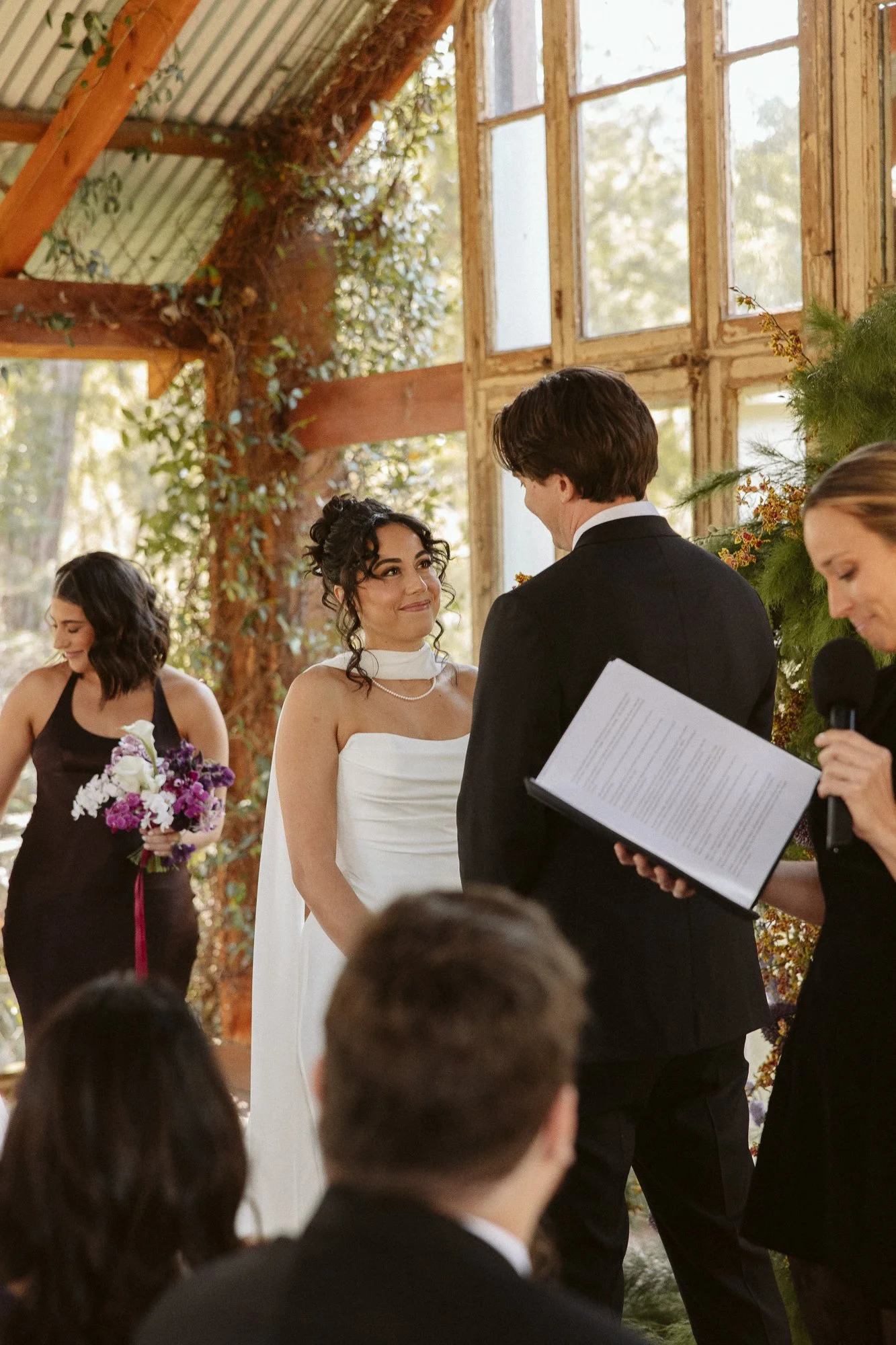 A wedding ceremony with a bride and groom exchanging vows inside a glass-roofed, wood-framed greenhouse filled with greenery. The bride is wearing a white strapless dress, and the groom is in a black suit. An officiant holds a book and is speaking in