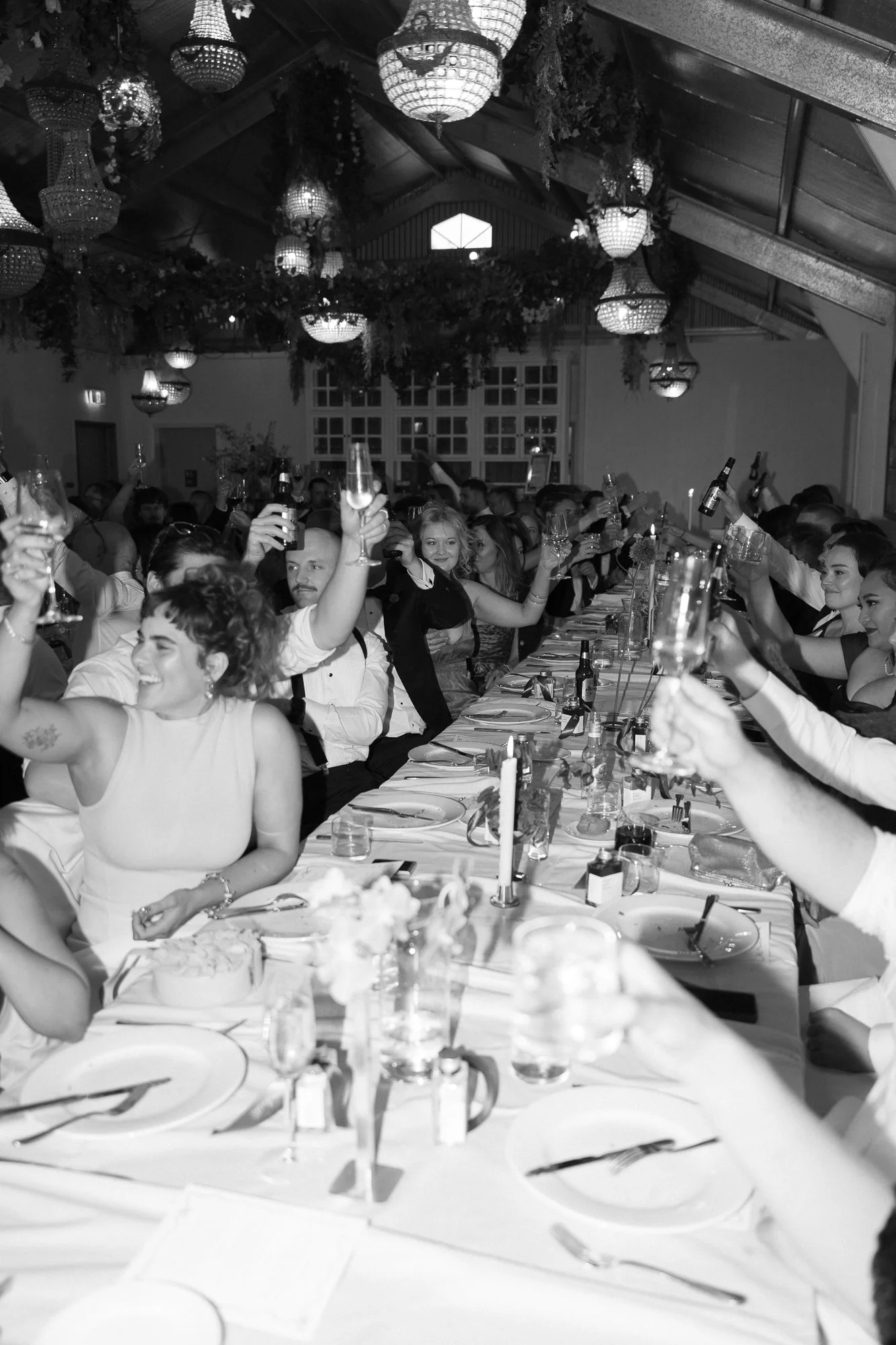 Black and white photo of a large group of people gathered at a banquet table, raising glasses in celebration during a dinner event, under decorative hanging lights.