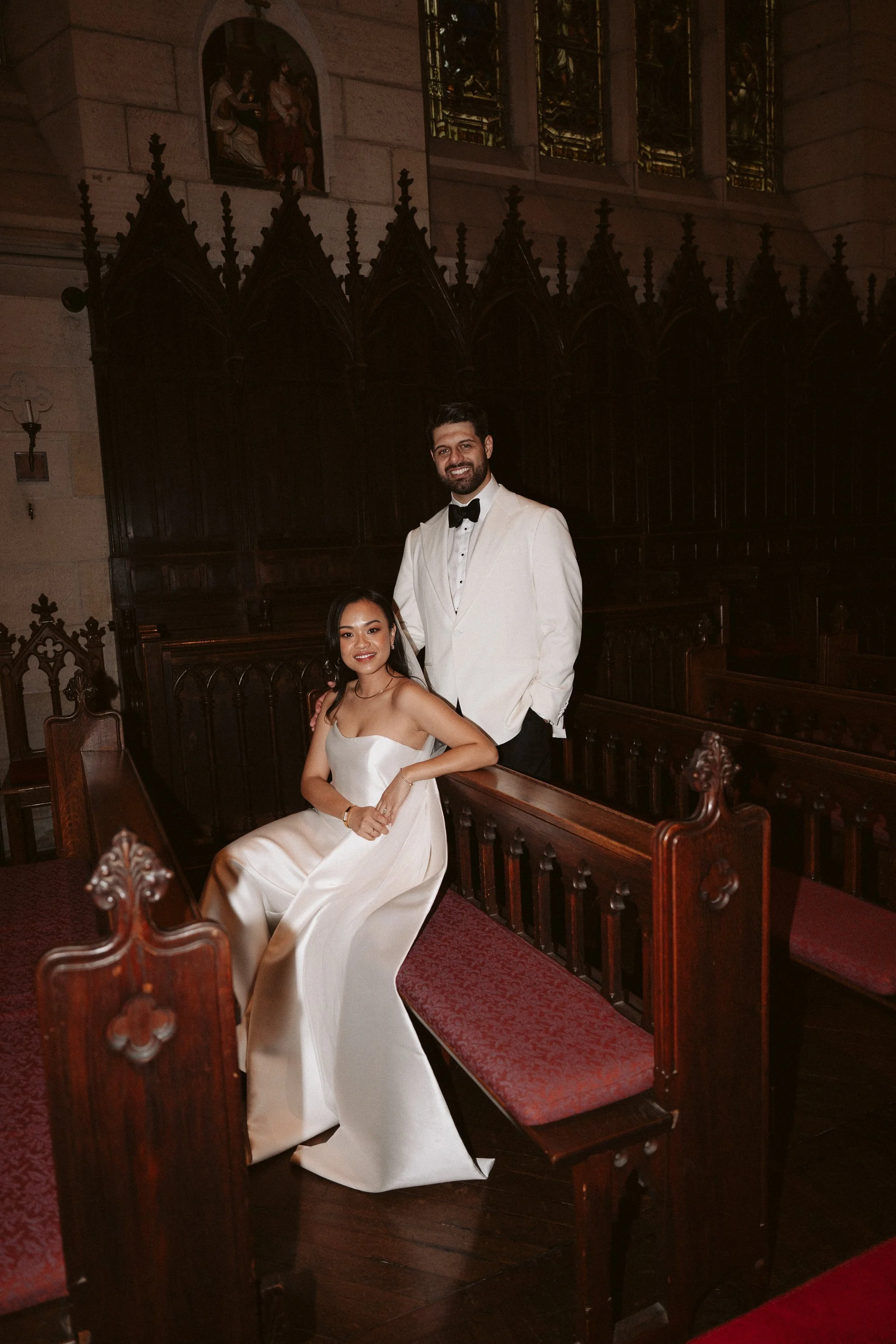 A man in a white tuxedo with a black bow tie standing, and a woman in a white dress sitting on a wooden pew inside a church with stained glass windows.