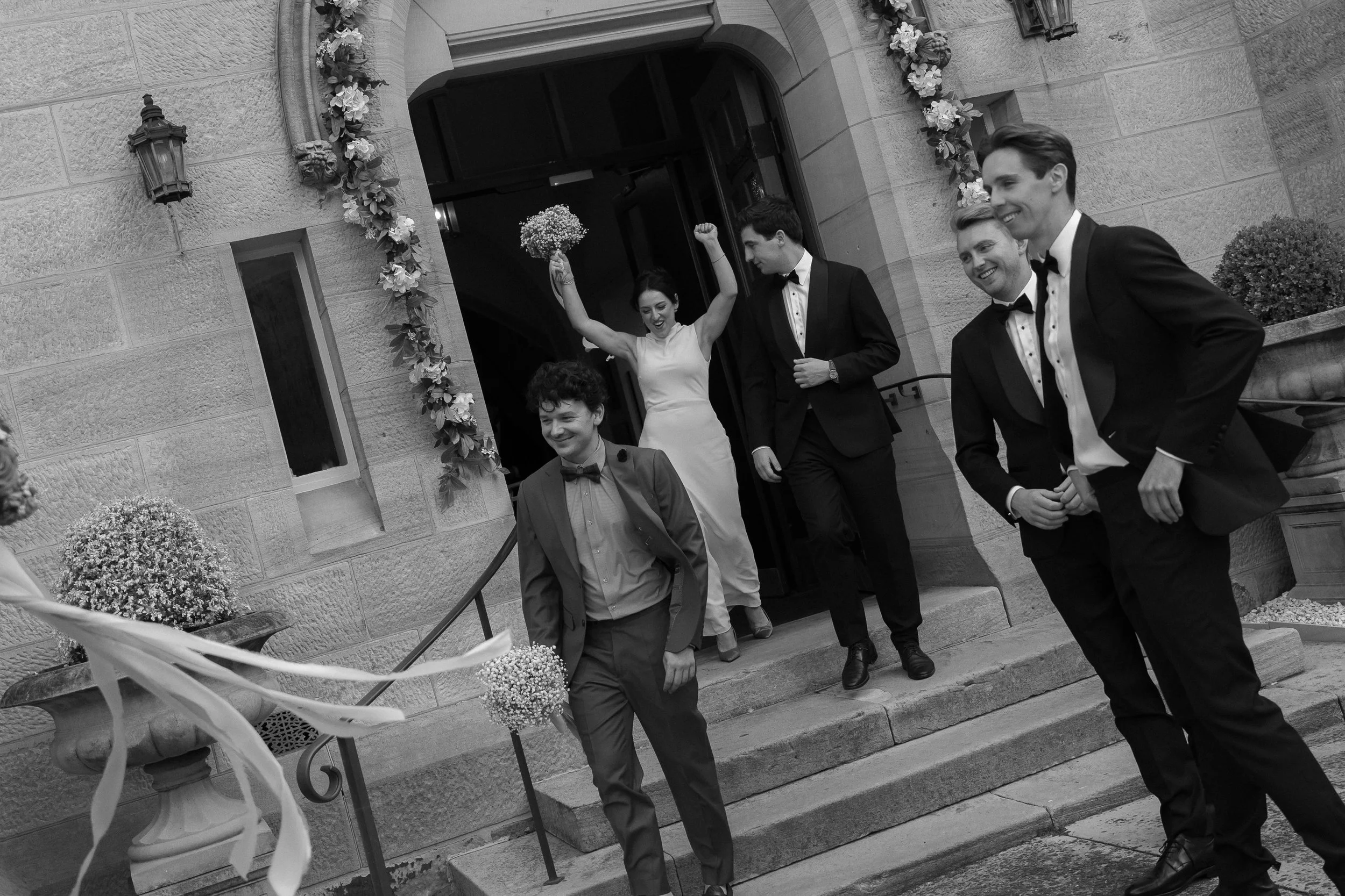 Celebrating group of people exiting a building decorated with flowers for a wedding, with happy expressions and a bride holding a bouquet up in the air.