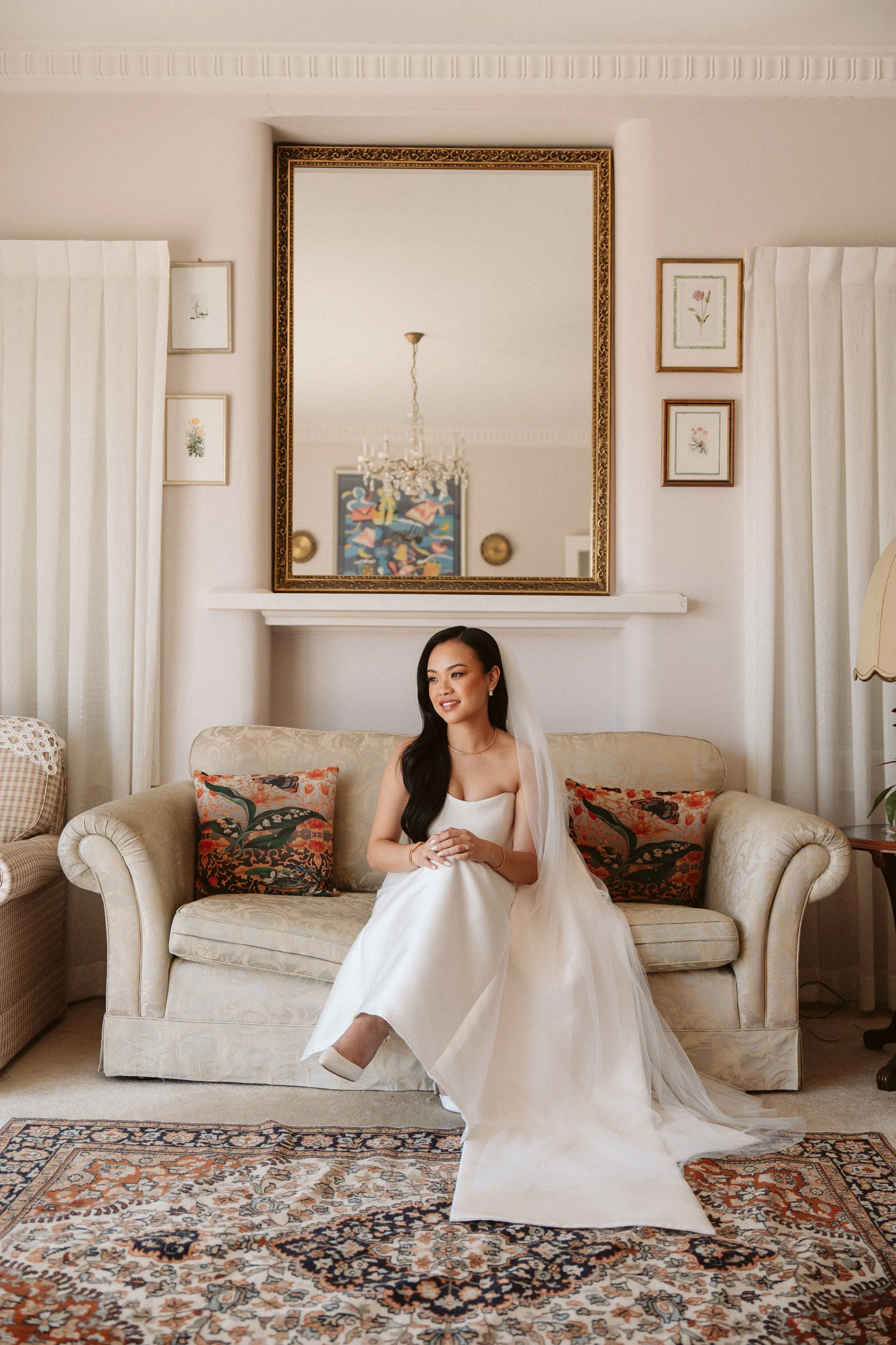 A bride in a white wedding gown sitting on a beige sofa in a living room with a large mirror above, framed pictures on the wall, and a patterned rug on the floor.