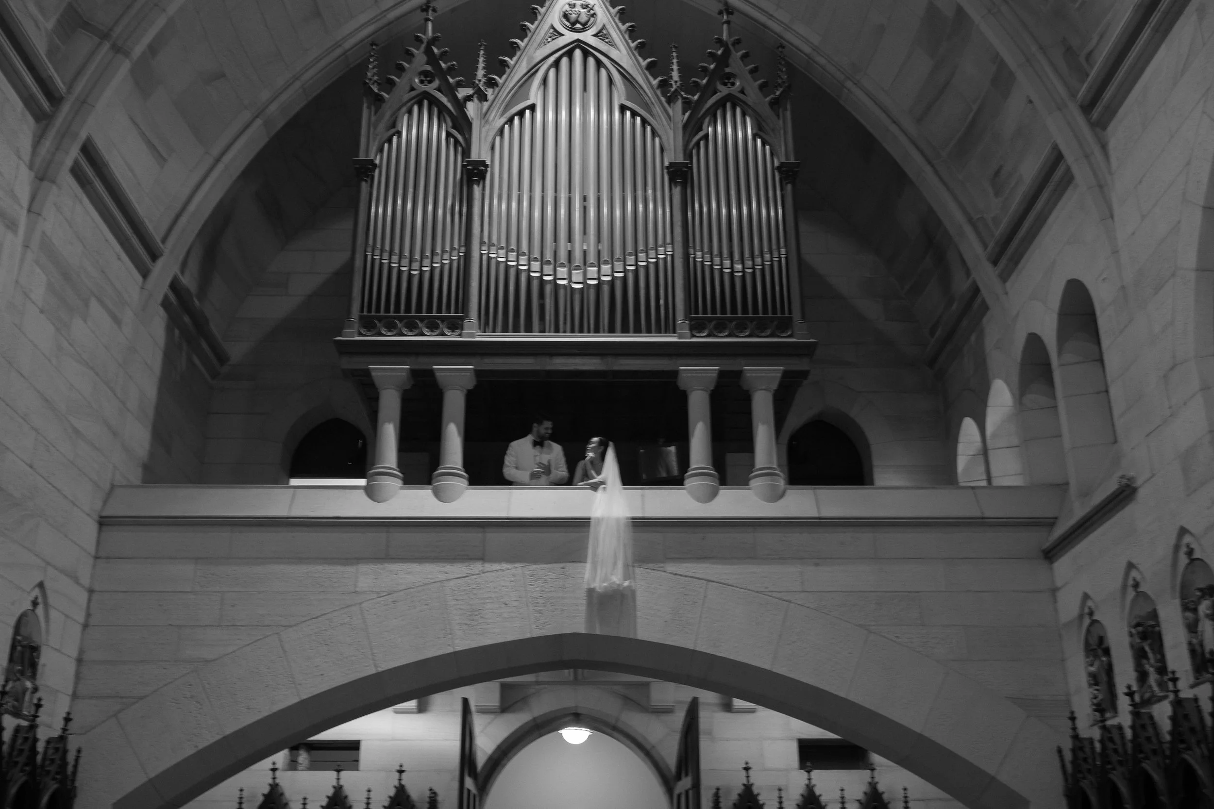 A black and white photo of a bride and groom standing in front of a large pipe organ in a church. The bride is wearing a wedding dress with a veil, and the groom is in a suit. The church has stone walls and arched windows.