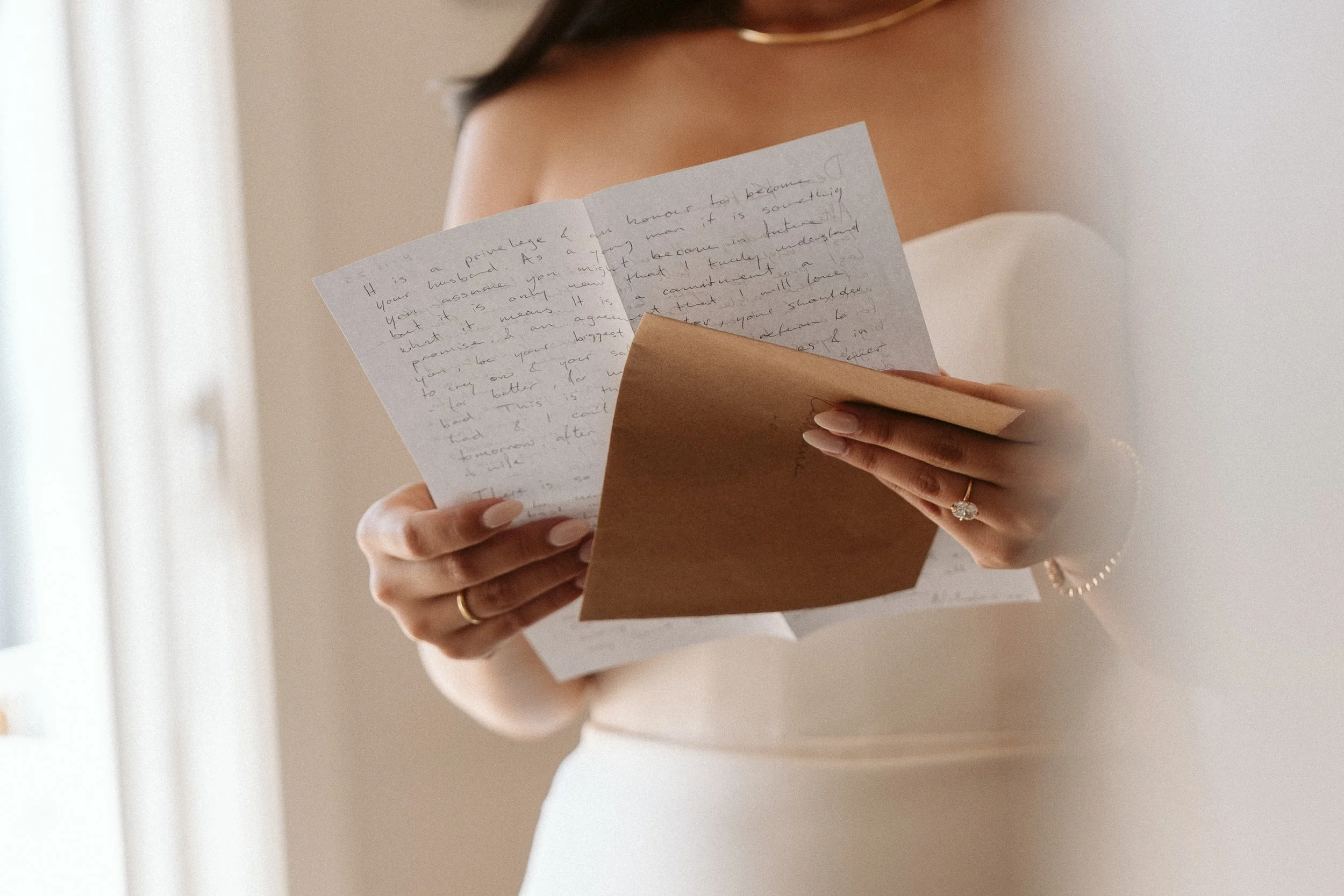 A woman in a white dress reading a handwritten letter and card, wearing rings and a pearl bracelet, with a blurred background.
