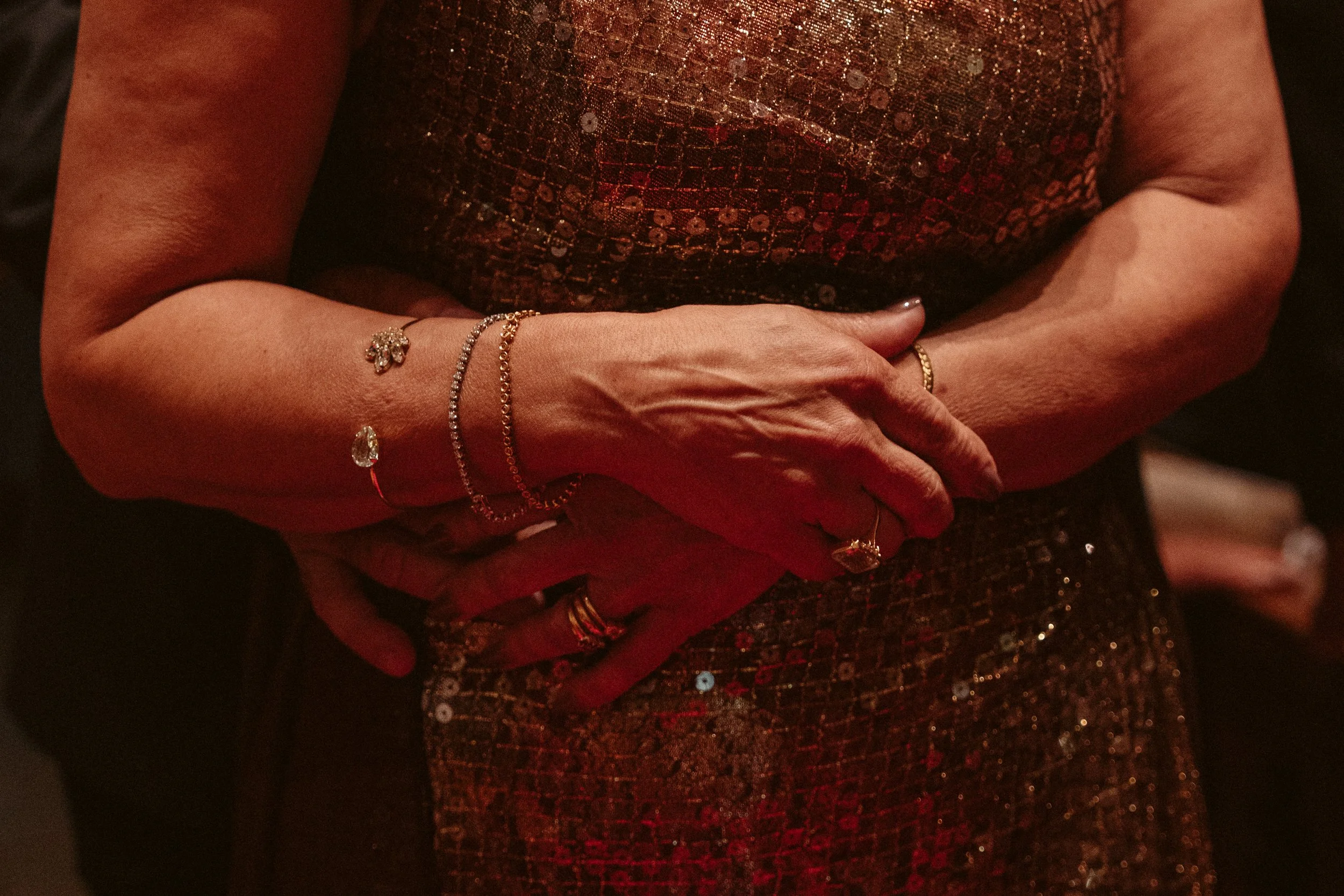 Close-up of a woman's hands with jewelry, wearing a sparkly dress in a dimly lit setting.