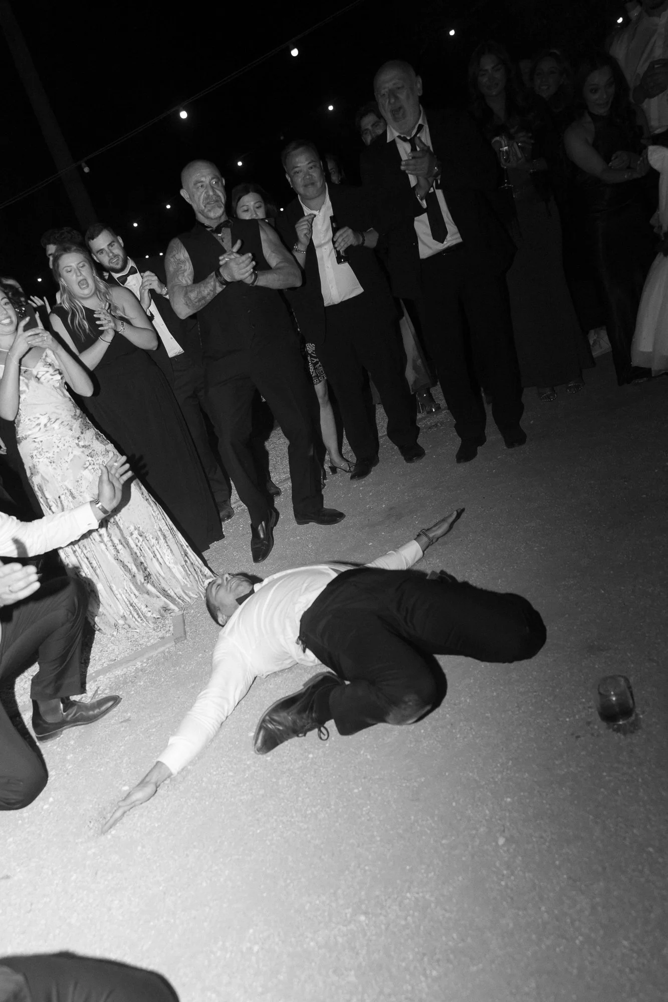 Man dressed in formal attire lying on the ground at a party or celebration, surrounded by standing guests in formal clothing, some holding drinks, with string lights above.