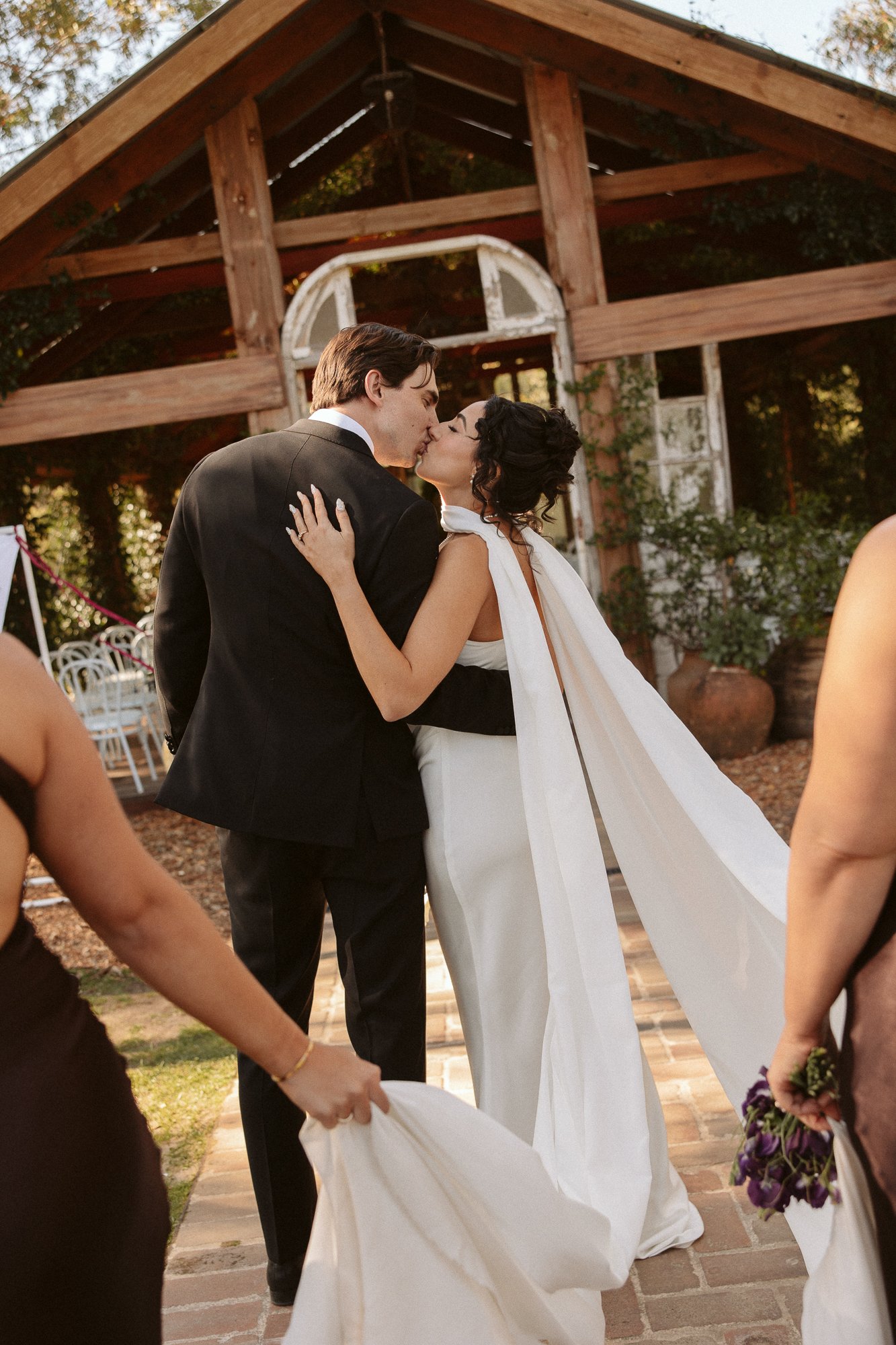 A newlywed couple sharing a kiss during their outdoor wedding ceremony, surrounded by friends and family, with a rustic wooden building in the background.