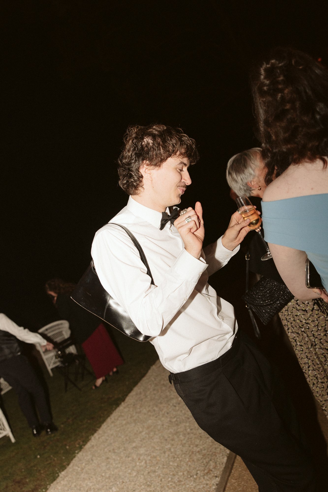 A young man in formal attire dancing at a party or event at night while holding a drink in his hand.