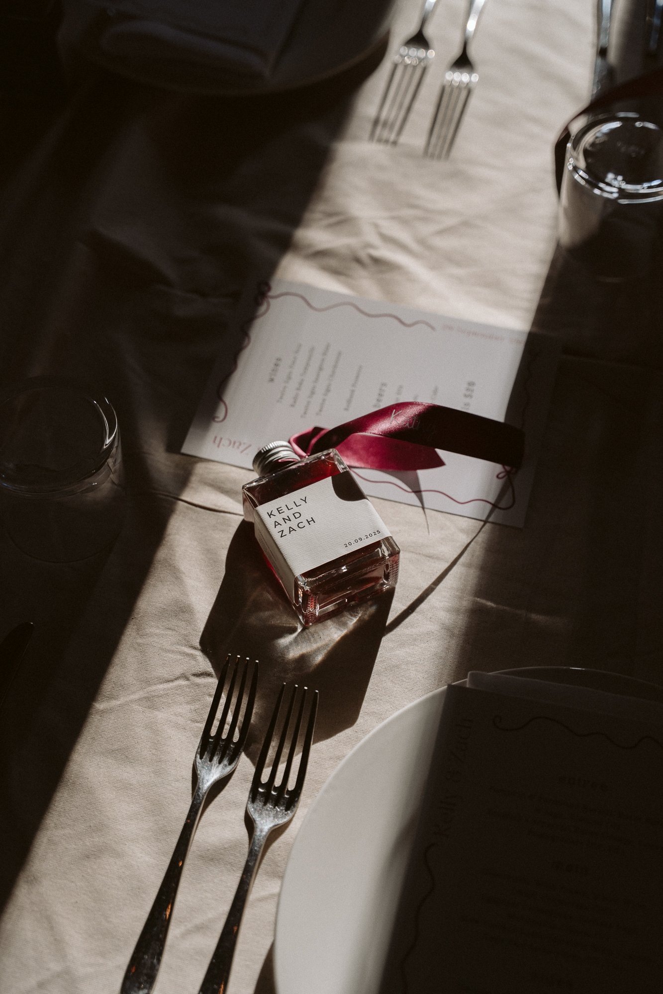 A table setting with a small glass bottle with a pink ribbon, labeled with the names Kelly and Zach and a date, possibly a wedding favor. The table has utensils, glasses, a menu, and a partially visible white plate, with sunlight casting shadows.