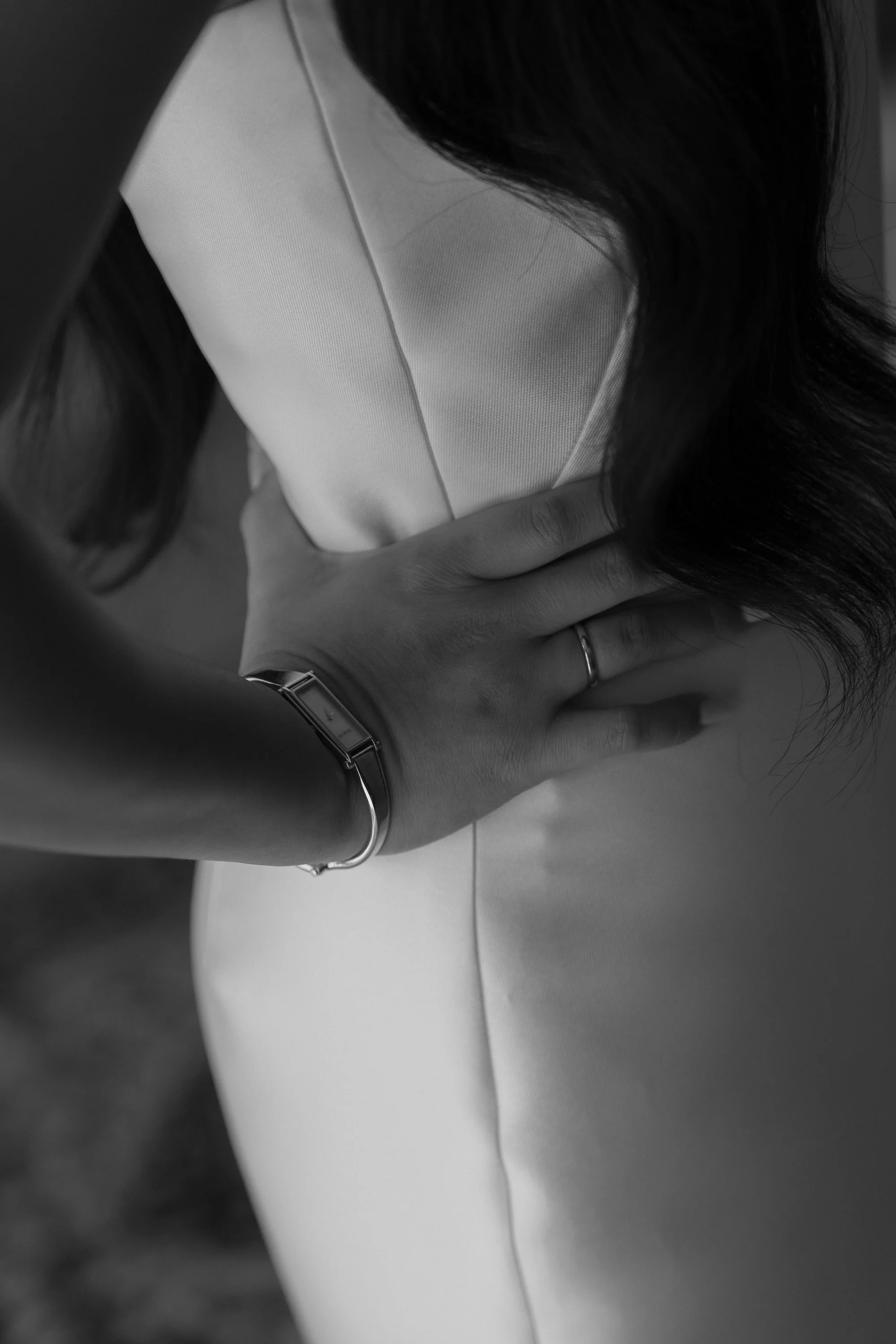 A person with long dark hair wearing a wristwatch and a ring, resting their hand on a light-colored cushion or piece of furniture.