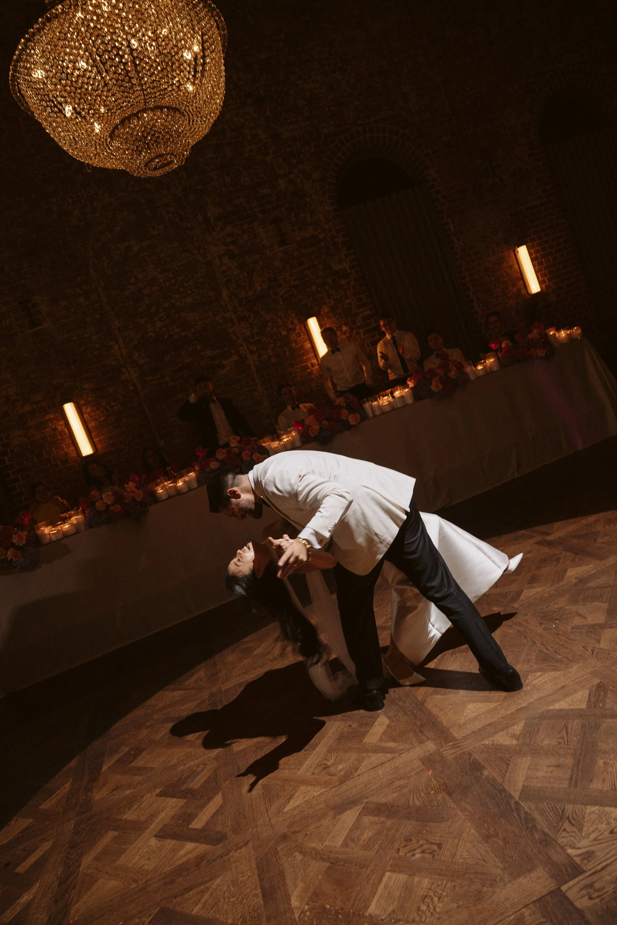 People dancing at a wedding reception in a dimly lit, elegant venue with a large chandelier and floral decorations on the tables.