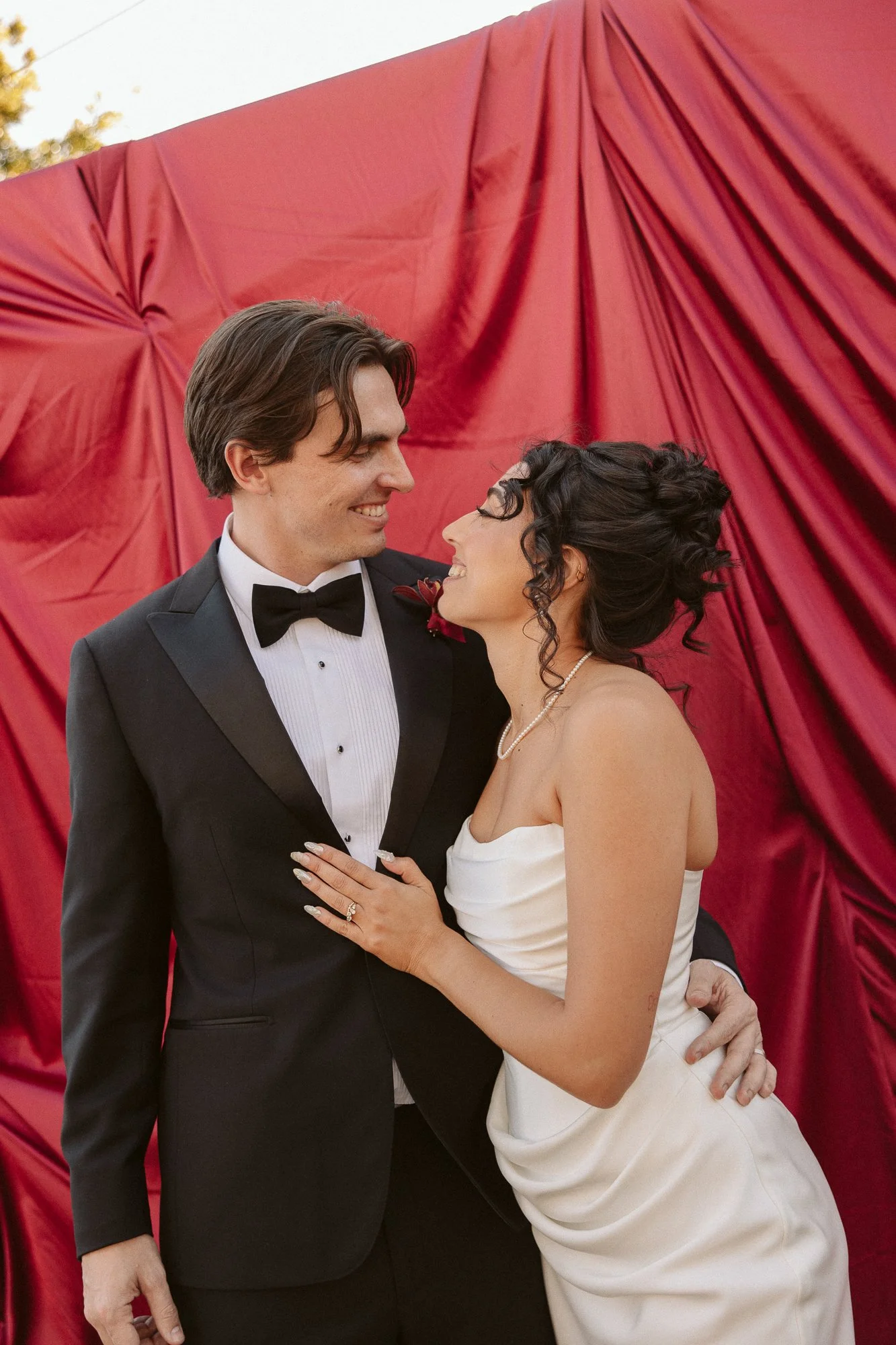 A newly married couple smiling at each other, standing in front of a red fabric backdrop, dressed in wedding attire.