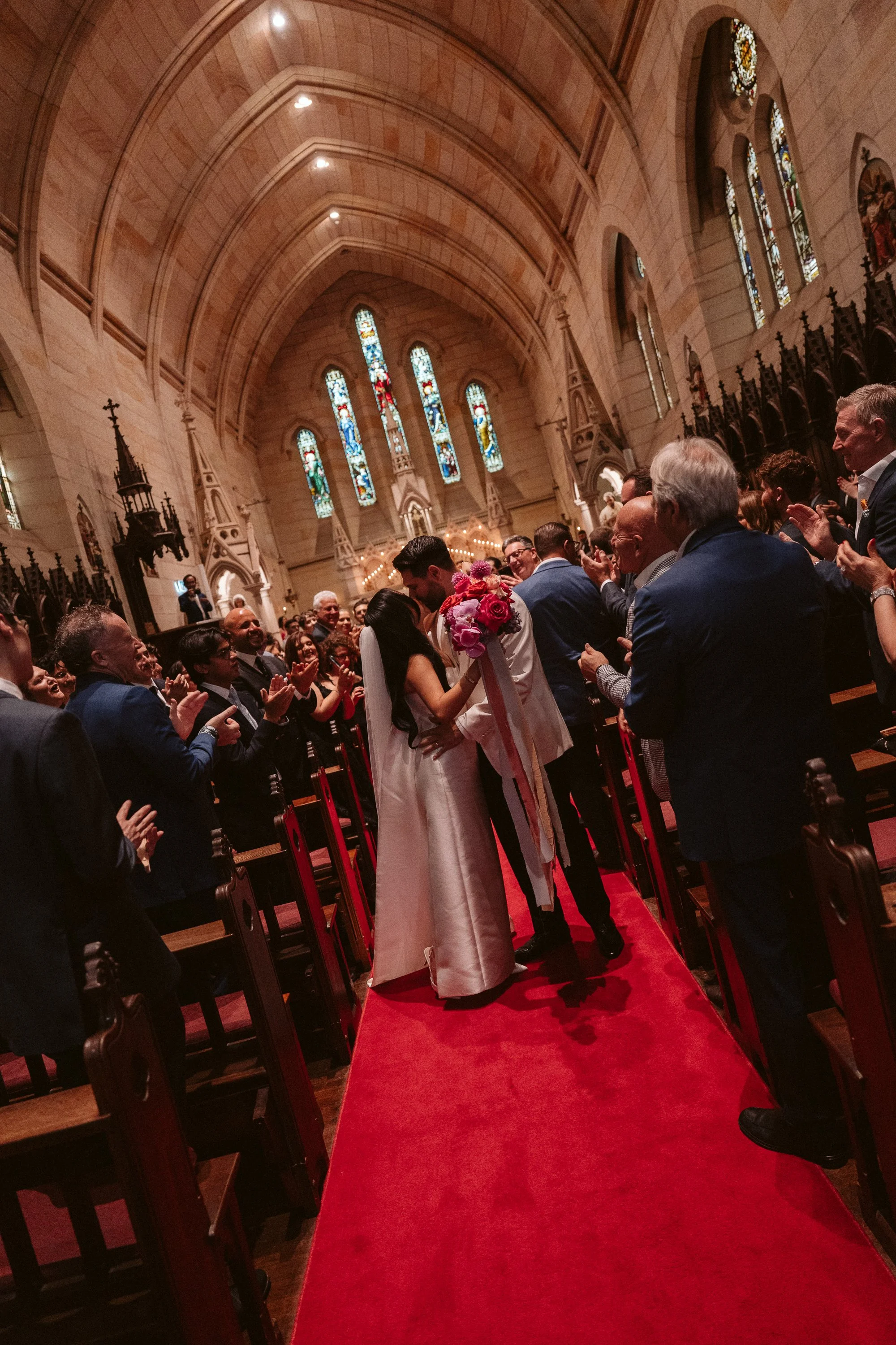 A wedding ceremony inside a grand church with stained glass windows. The bride and groom are kissing in front of seated guests who are applauding.