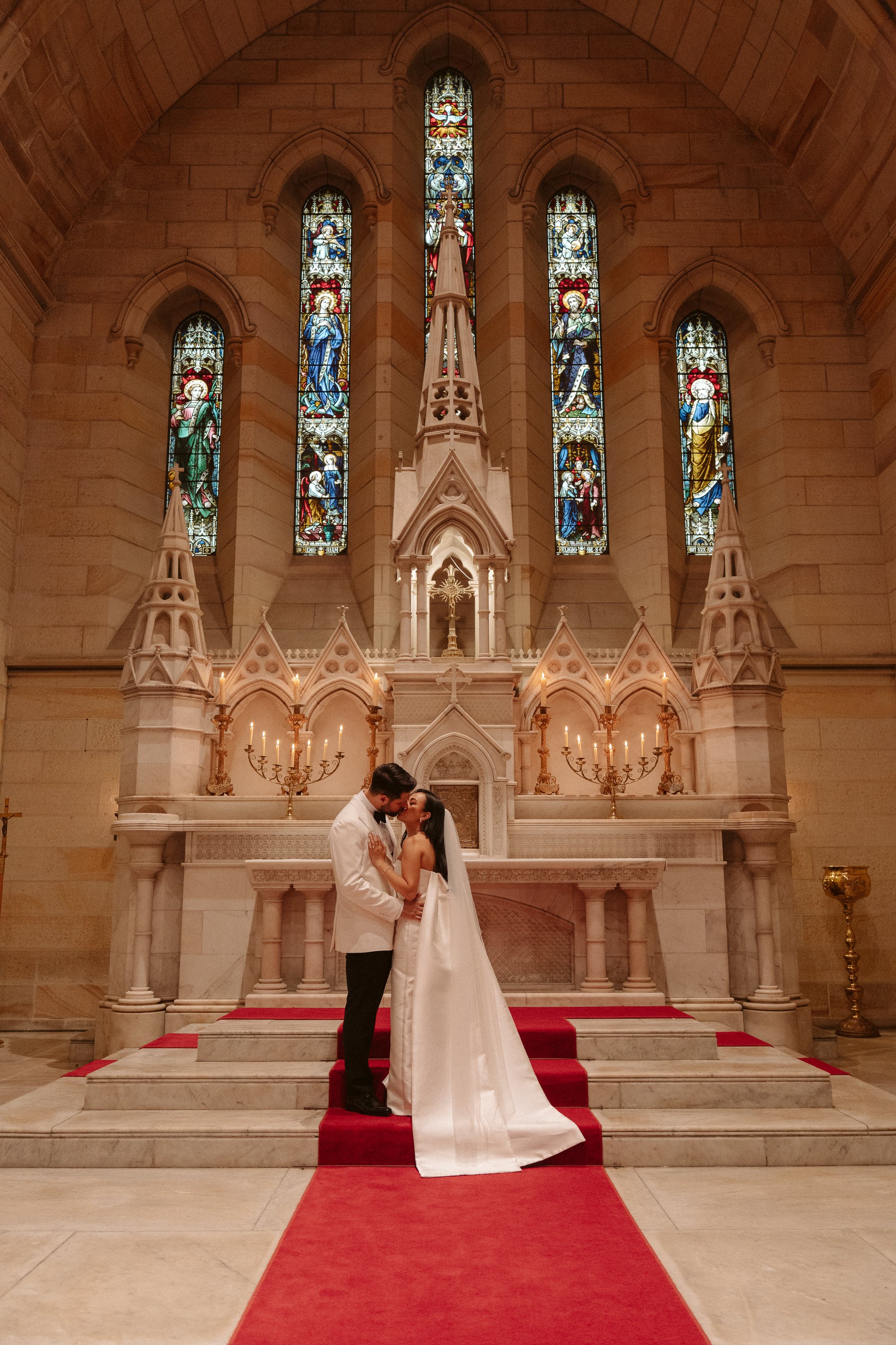 A bride and groom kissing at the altar inside a church, with stained glass windows and candle holders behind them.