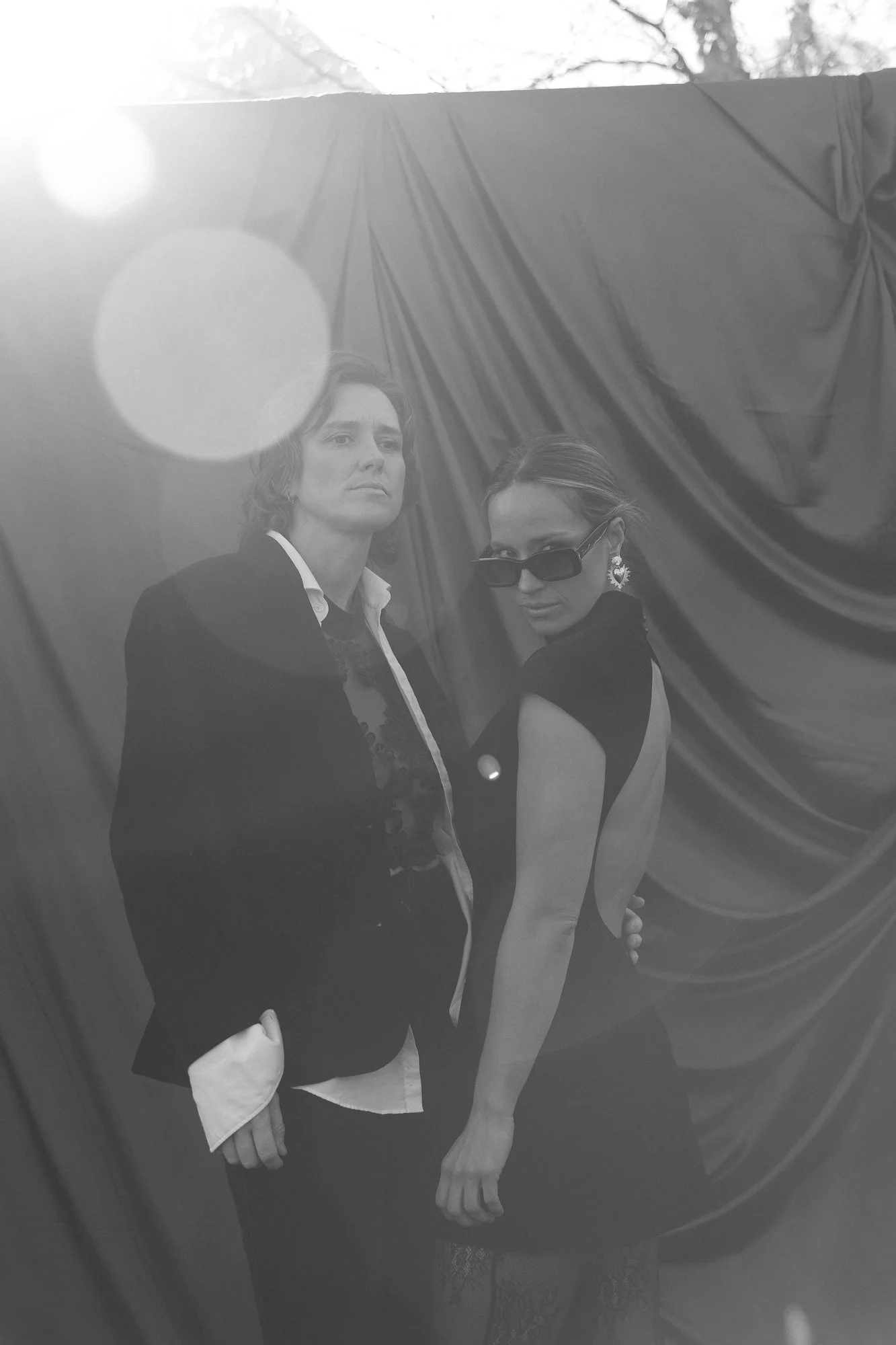 Black and white photo of two women standing in front of a fabric background with lens flare effects. One woman is wearing sunglasses and earrings, and the other has a serious expression.