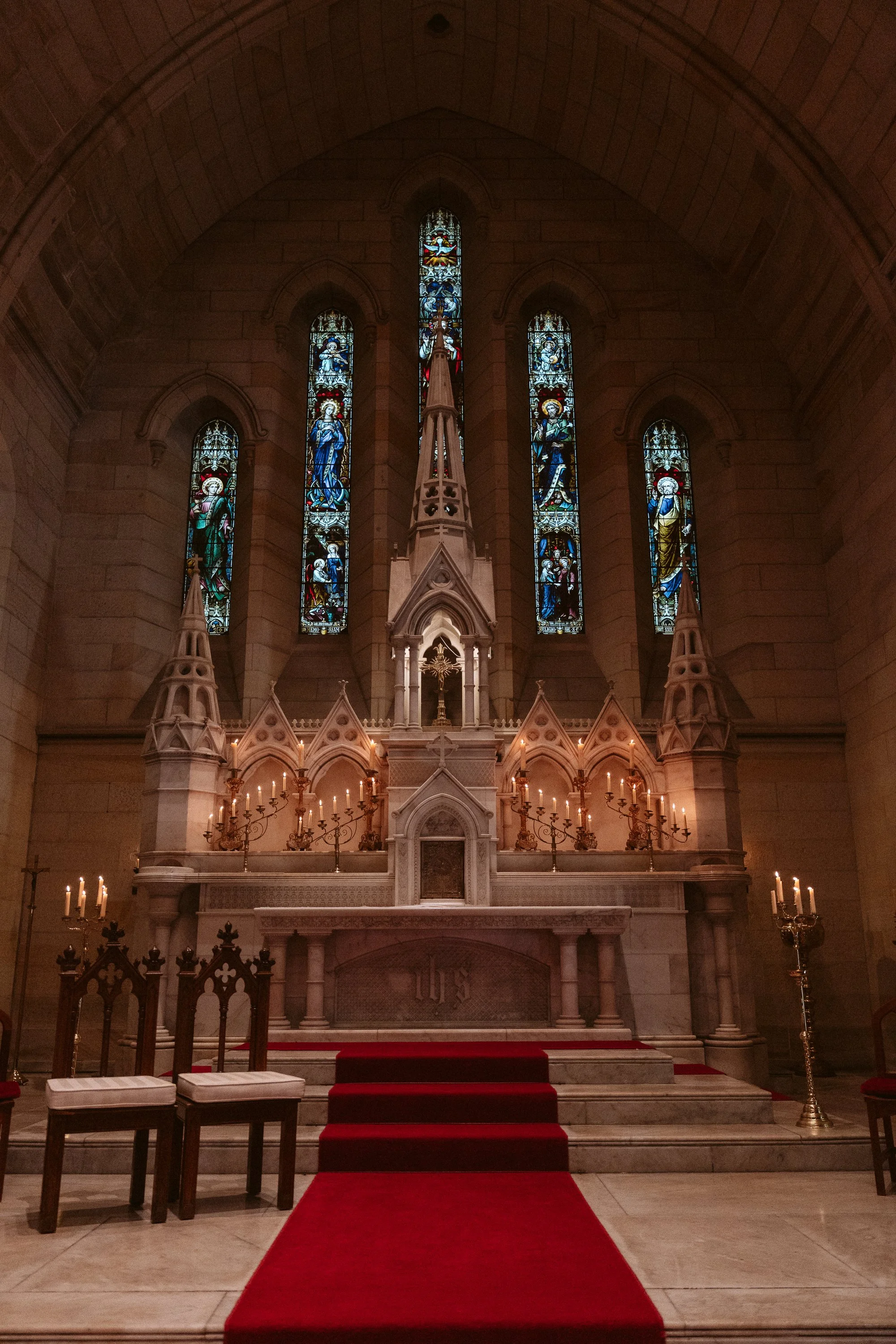 Inside a church, an ornate altar with candles and Gothic architecture, behind the altar are stained glass windows depicting religious figures.