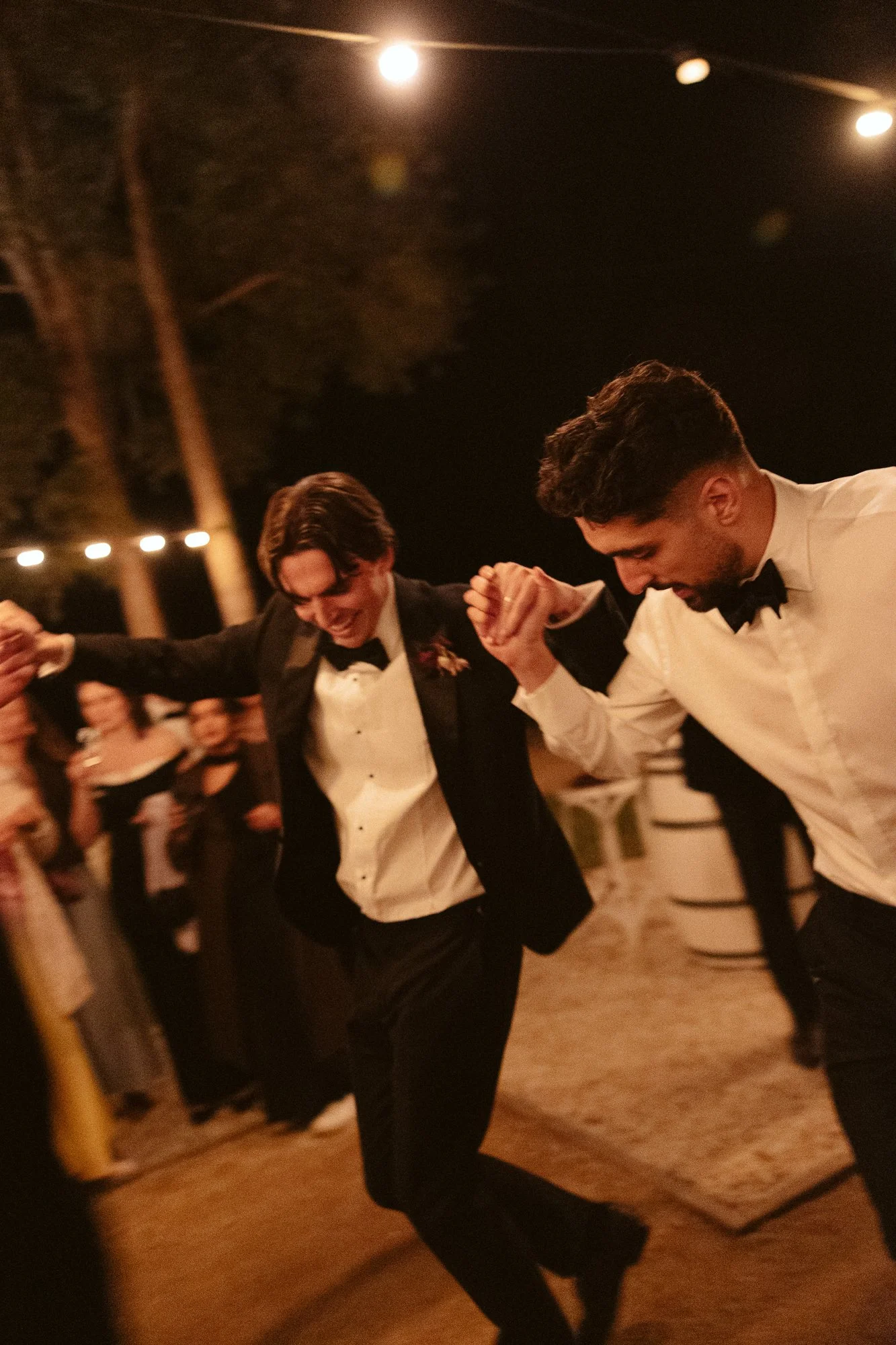 Two men in tuxedos dancing and holding hands at an outdoor celebration at night.