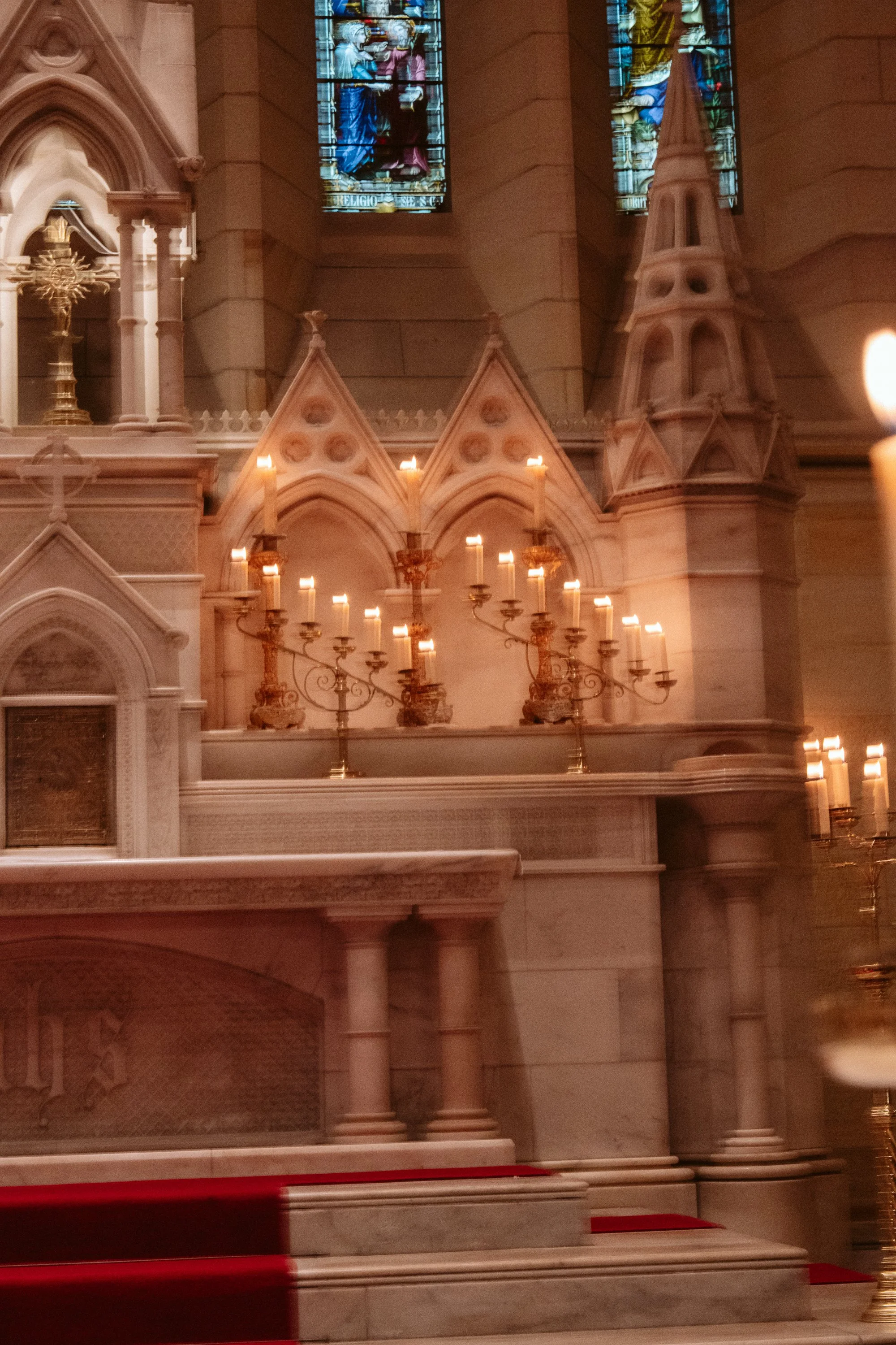 Interior of a church altar with multiple lit candles, intricate gothic architecture, stained glass windows depicting religious scenes.