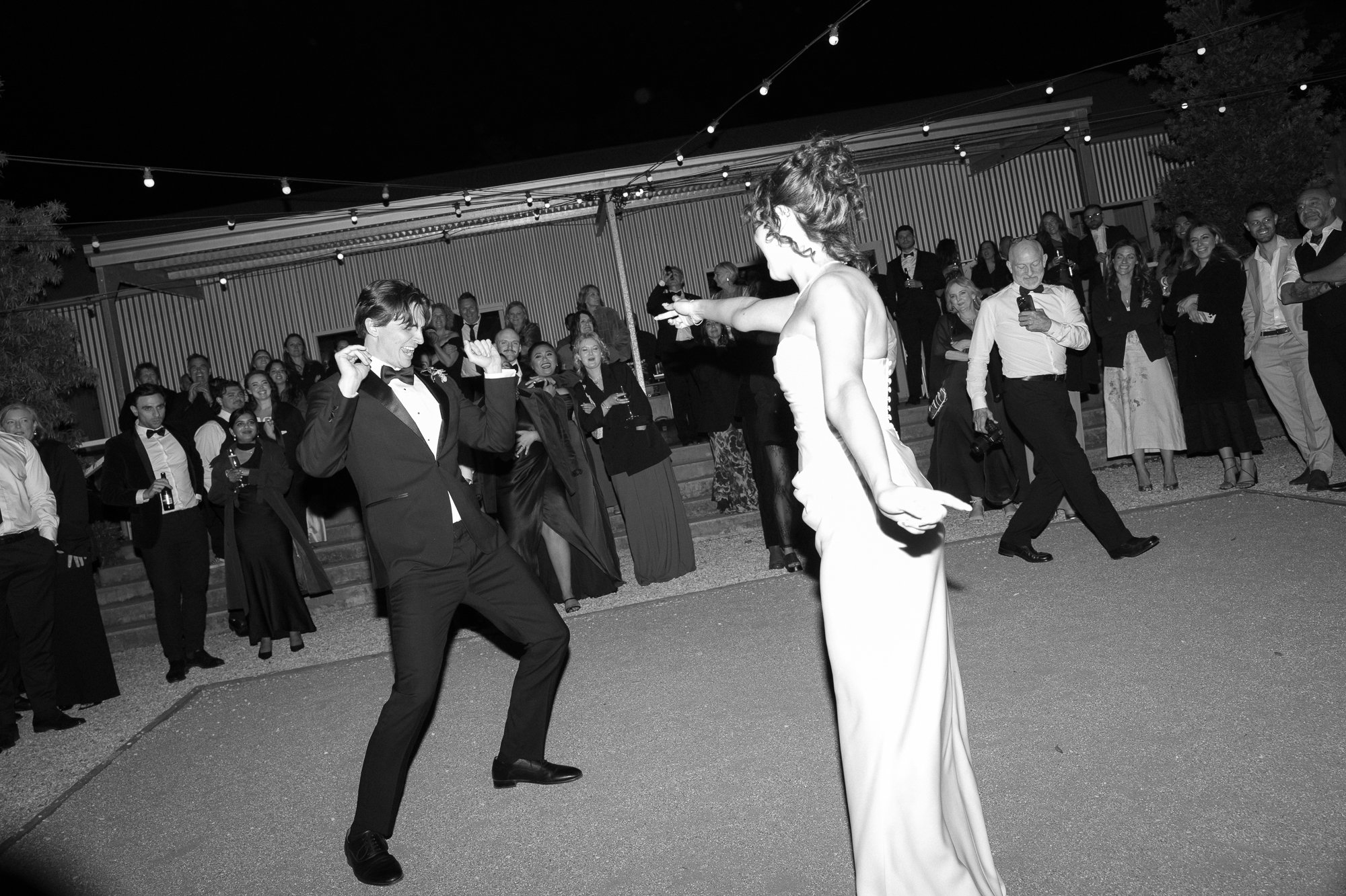 A woman in a strapless gown dancing with a man in a tuxedo at an outdoor party at night, with onlookers in formal attire watching in the background.