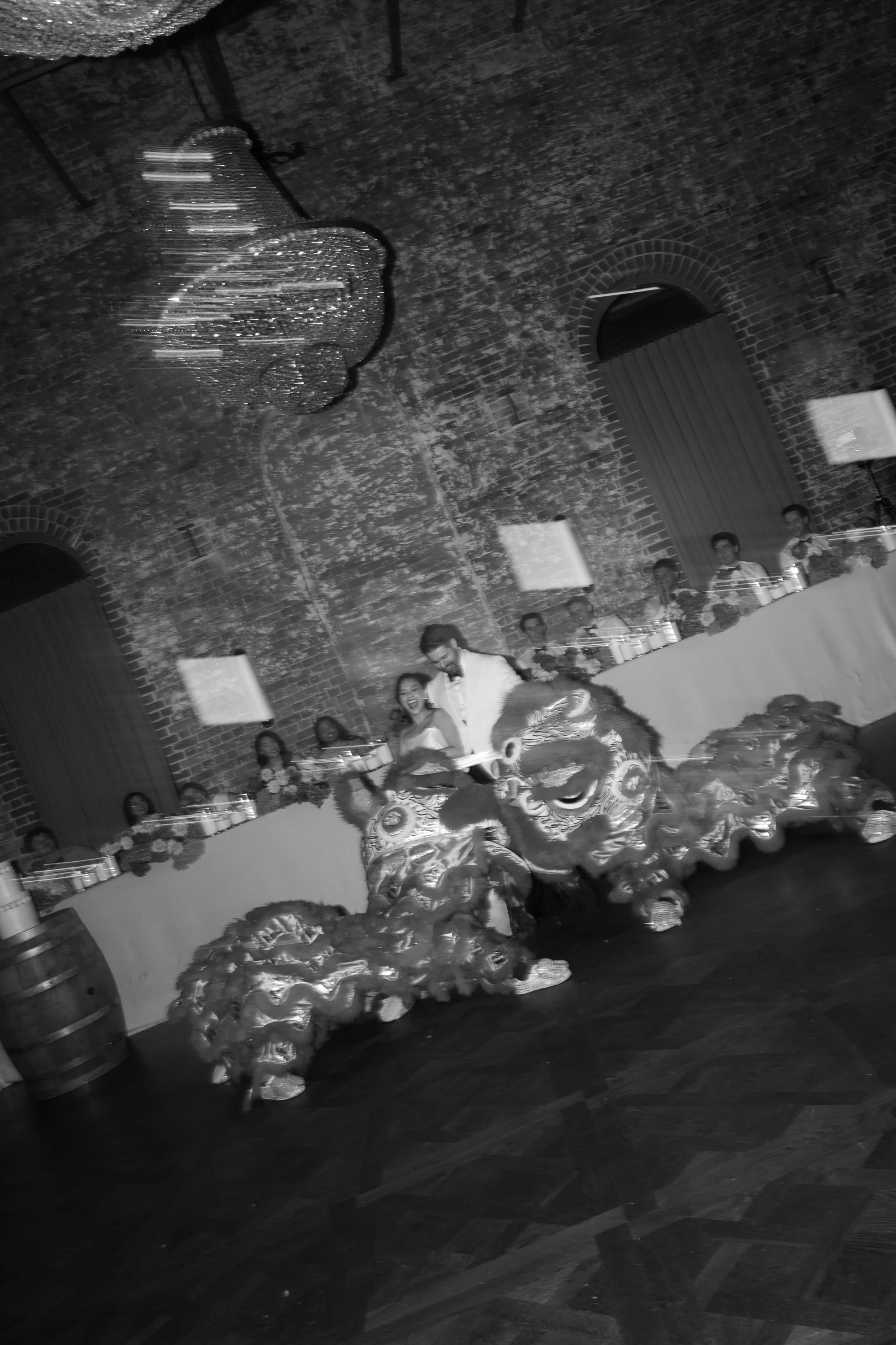 A couple riding a traditional Chinese lion dance costume during a celebration, with guests seated at long tables and a large chandelier hanging from an exposed brick wall ceiling.