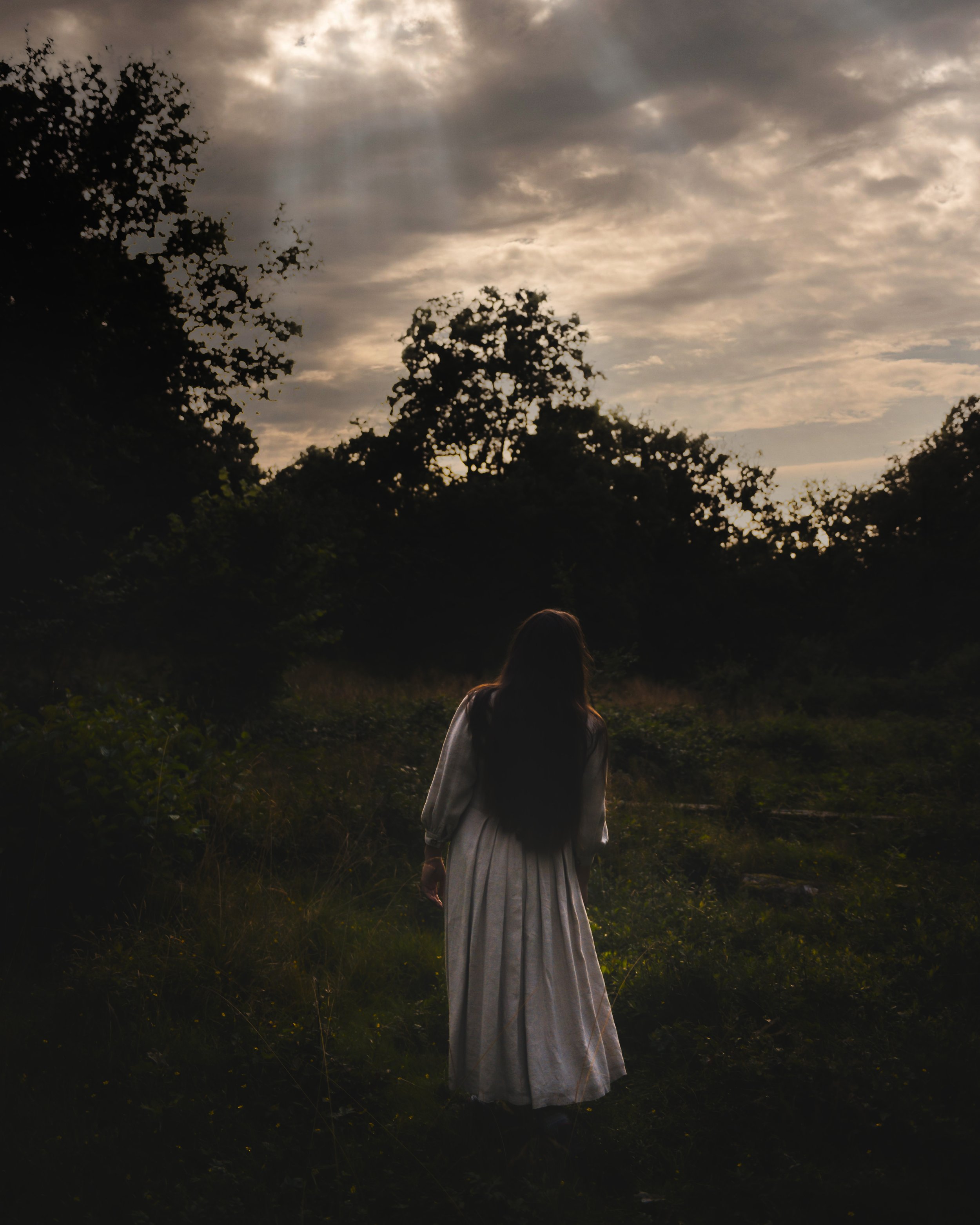 Woman in a white dress standing in a dimly lit woodland area with a cloudy sky overhead.
