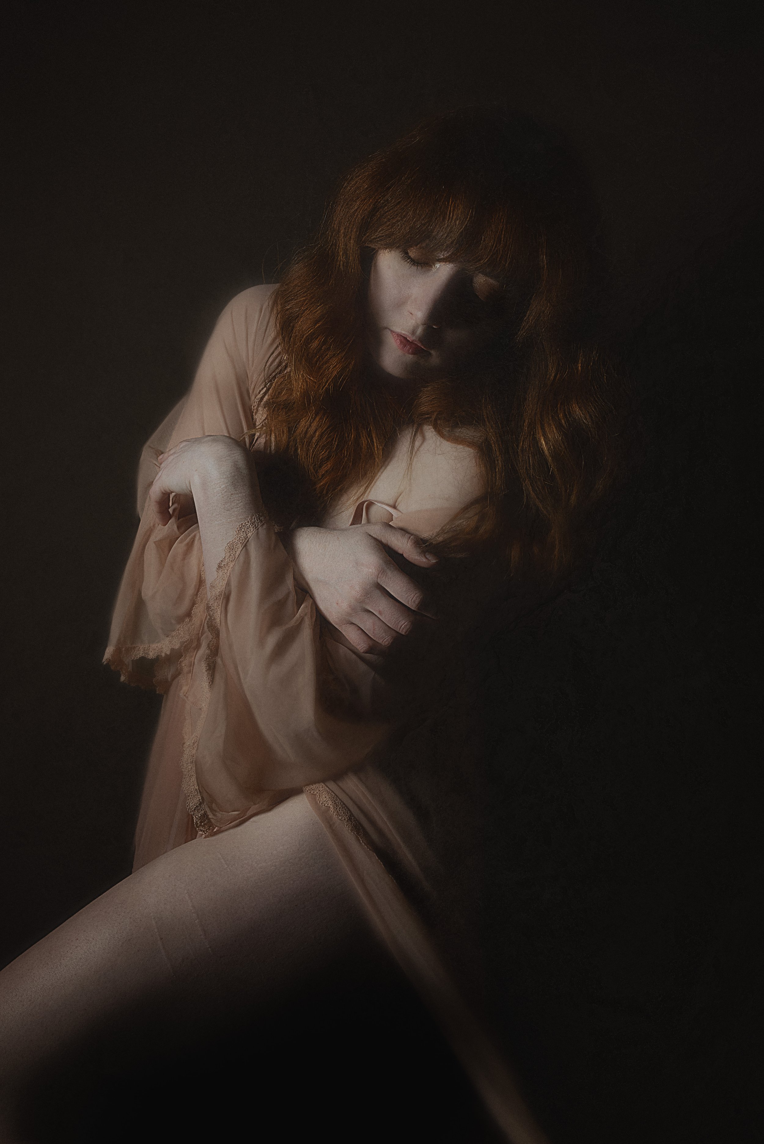 Portrait of a woman with long red hair, wearing a sheer garment, against a dark background. She is hugging herself and looking downward with a serene expression.