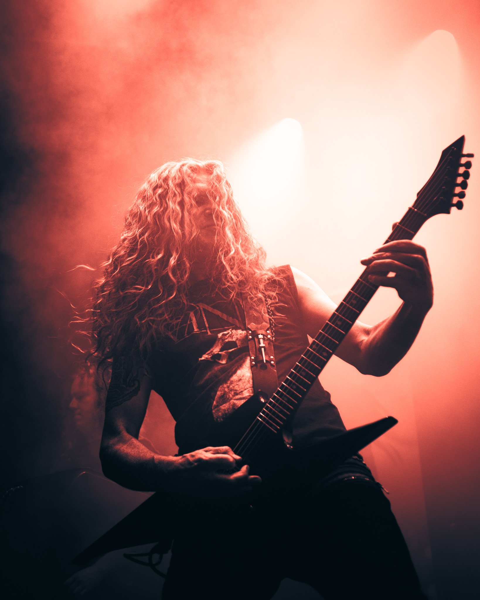 Guitarist with long hair playing an electric guitar on stage with red lighting.