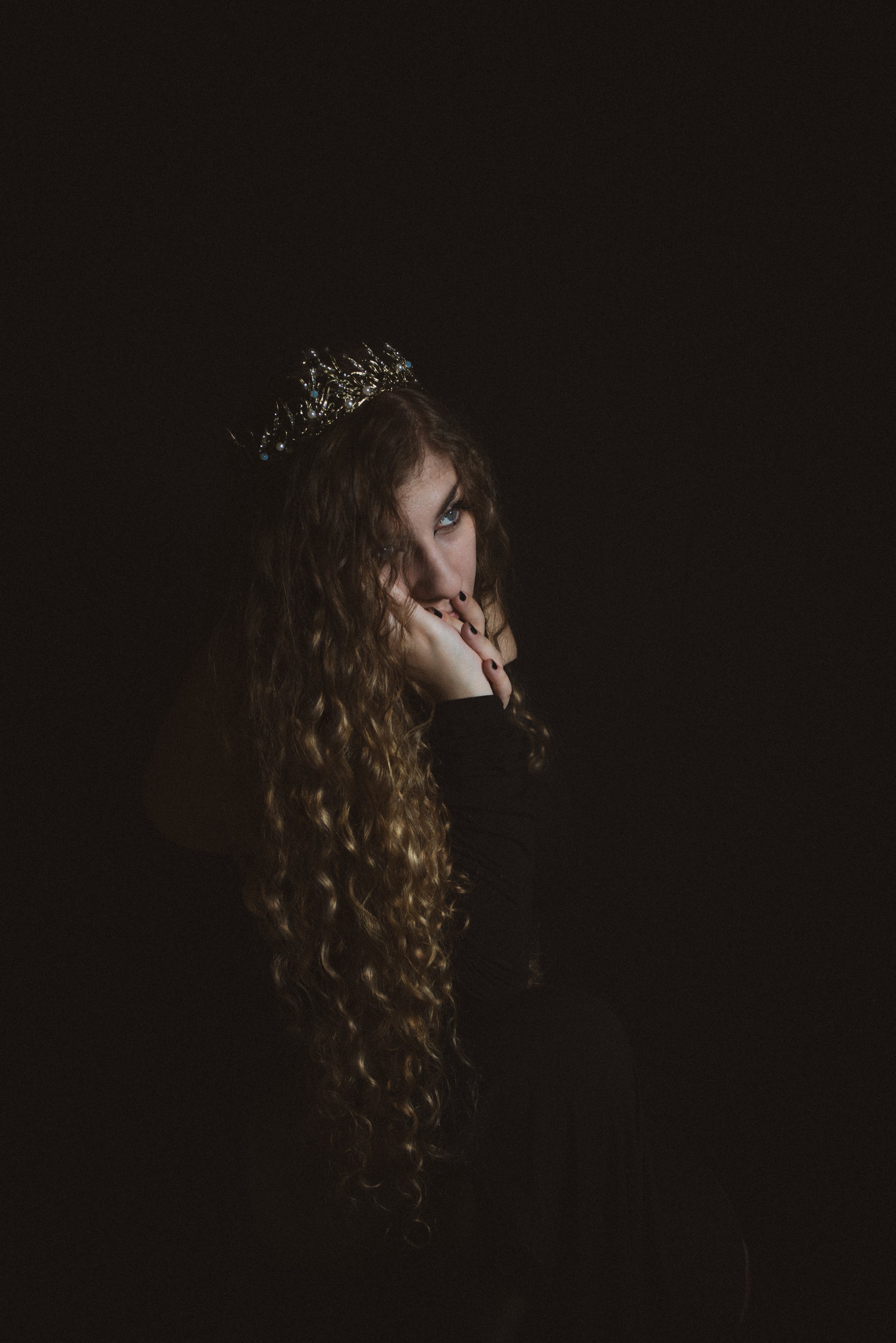 Person with curly hair and a crown sitting in a dark setting, with hand on face.